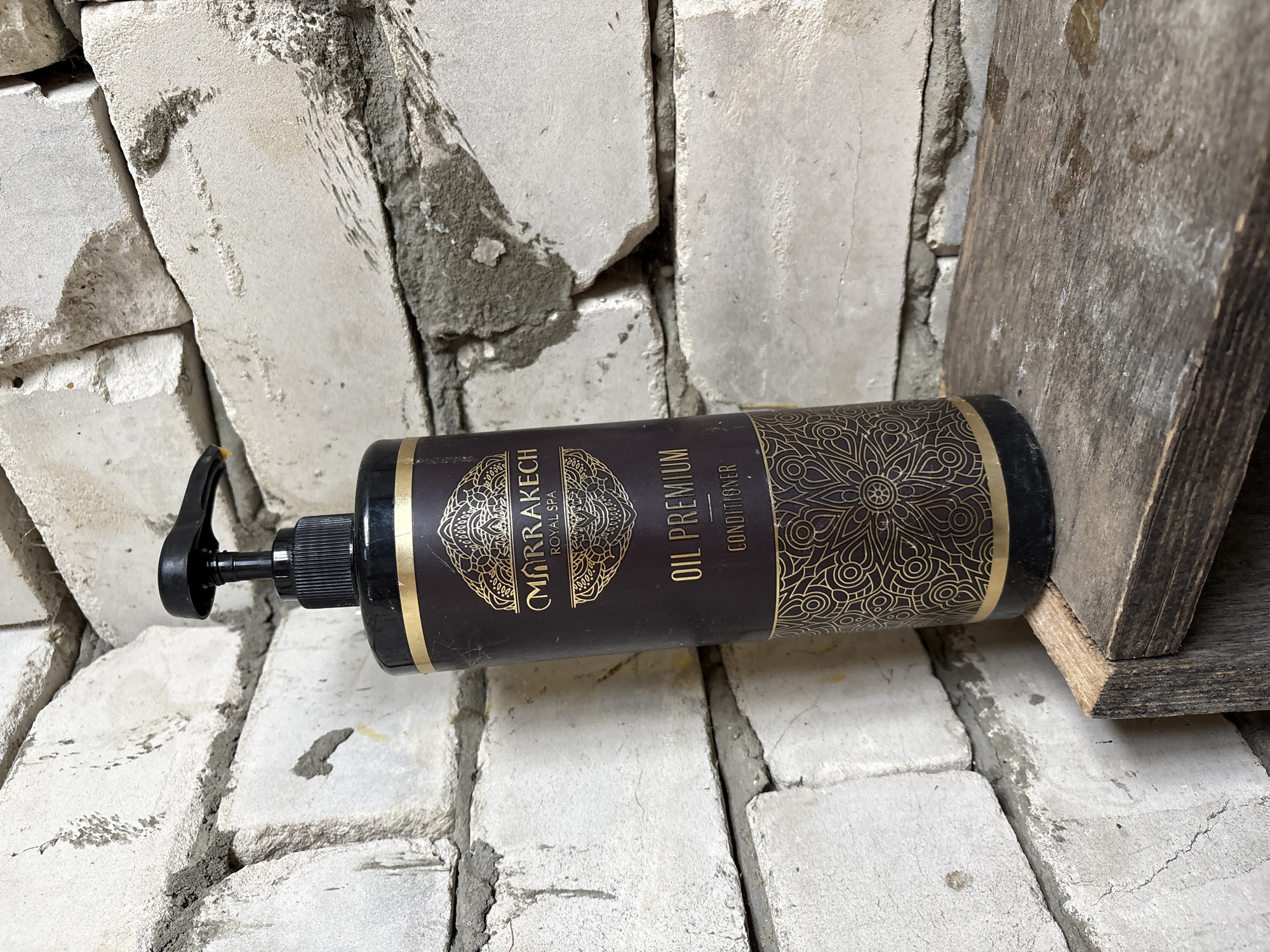 A dark brown bottle with a black pump dispenser stands on a rough wooden surface. The bottle features a gold emblem and text that reads "MARRAKECH ROYAL SPA OIL PREMIUM CONDITIONER". The background consists of a light-colored brick wall. The scene appears to be indoors, with no discernible city or specific location identifiable.
