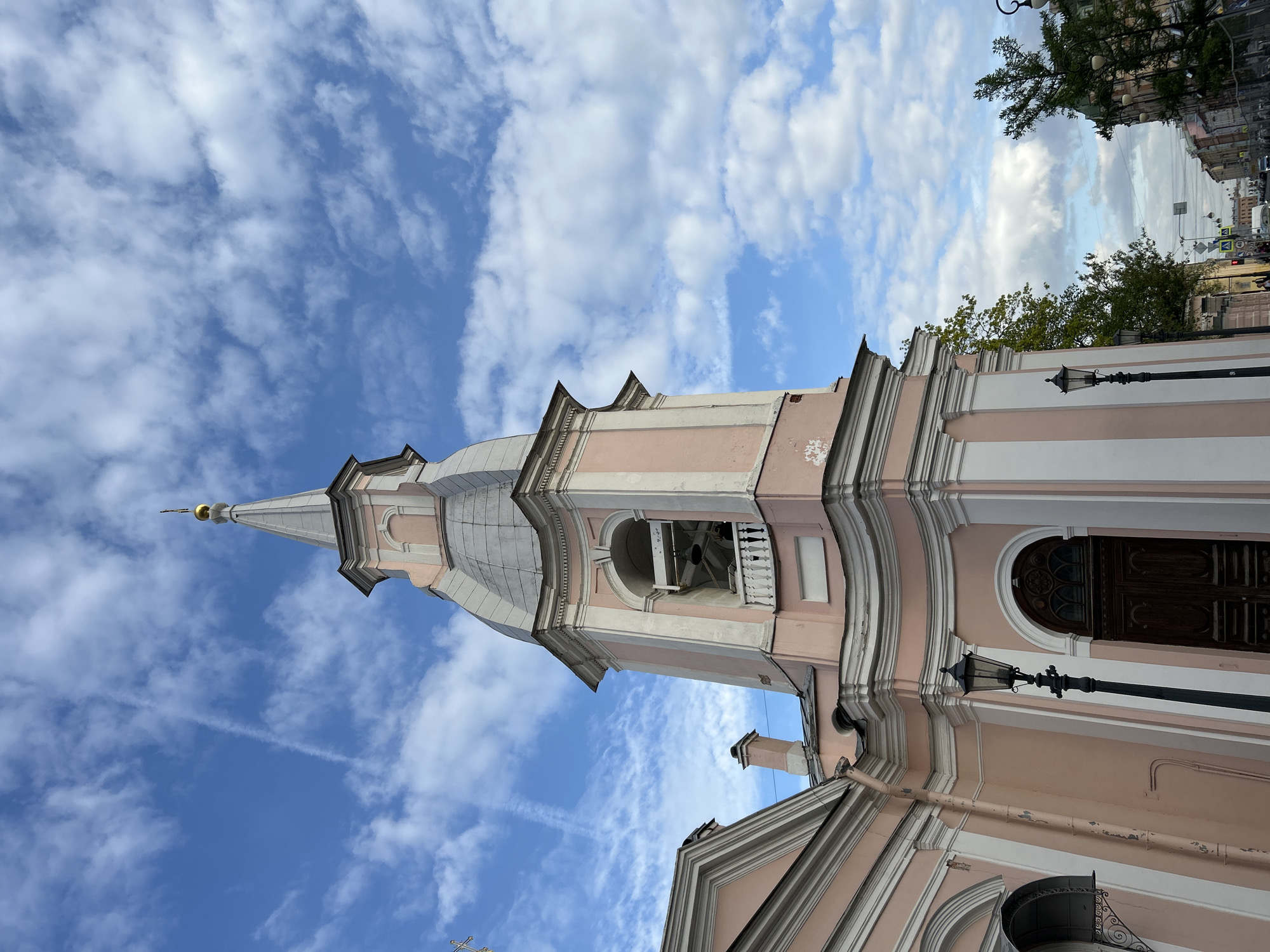 The image shows a low-angle view of the Church of Saints Simeon and Anna in Saint Petersburg, Russia. The church's pink and white facade is visible, along with its steeple rising into a blue sky filled with scattered clouds. A street with trees and buildings is partially visible to the right of the church. Several black street lamps are attached to the building.