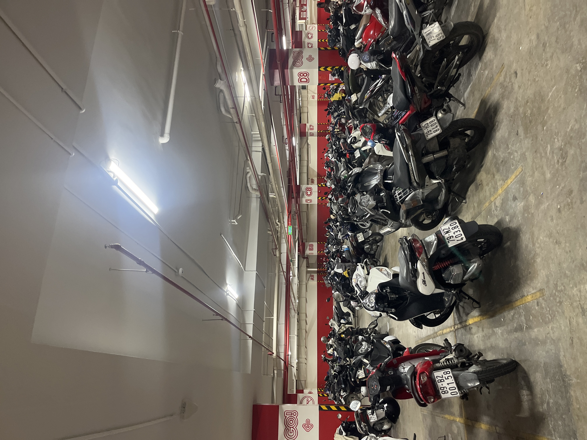 This is a medium-angle, eye-level, full shot of a crowded, multi-level underground parking garage in Tay Nha Trang Ward, Vietnam. The garage is filled with a dense arrangement of parked motorcycles, predominantly scooters. The concrete floor is marked with yellow lines indicating parking spaces. Red and white signage is visible throughout the garage, including "GO!" logos, directional arrows, and alphanumeric zone designations like "A8," "B8," "C7," and "D8." Pipes and conduits are suspended from the white ceiling, illuminated by fluorescent lights. The overall impression is one of an organized but heavily utilized vehicle storage facility. There are no people clearly visible in the image.