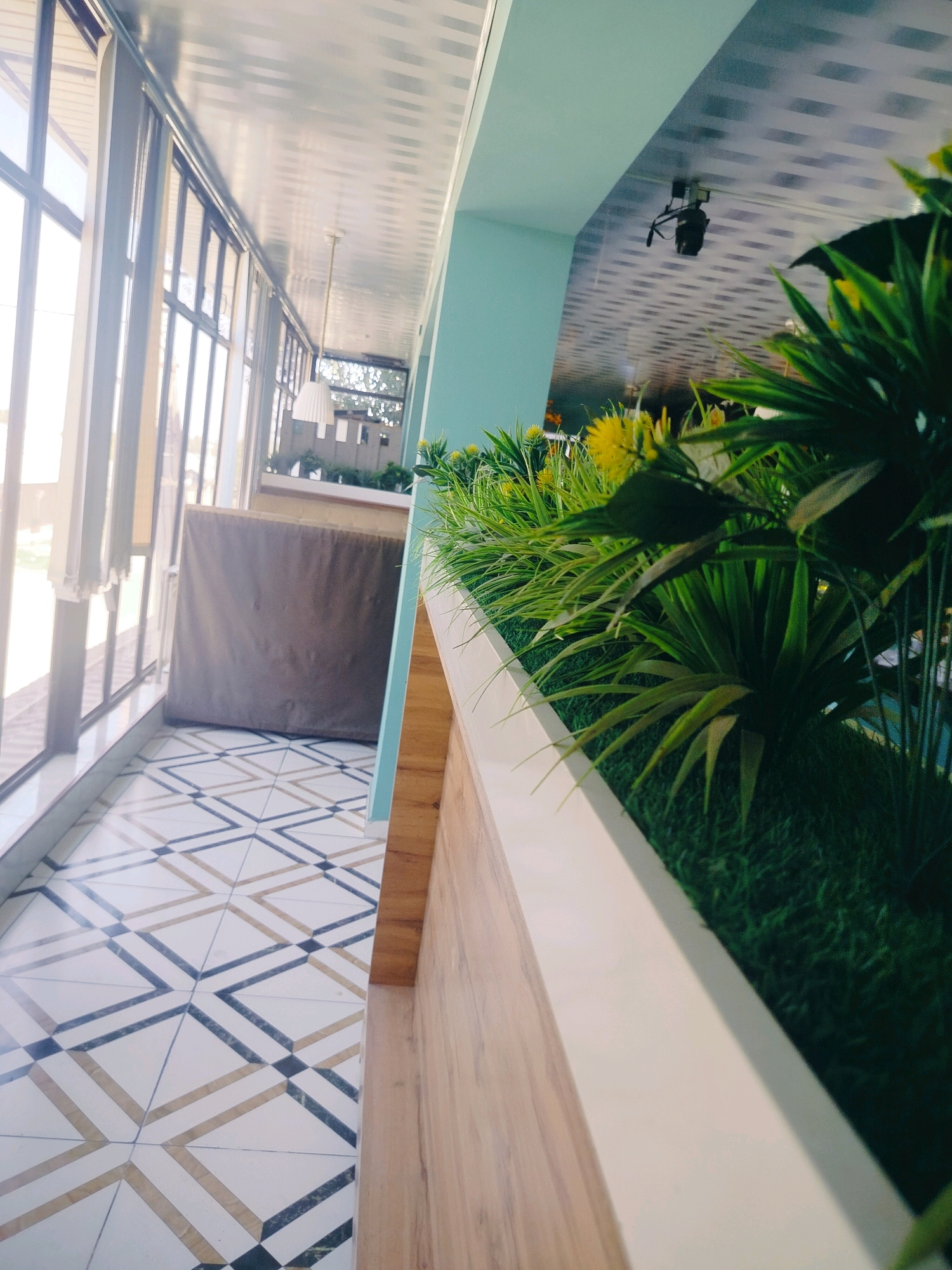 The image depicts an interior view of a modern establishment, possibly a cafe or restaurant, in the city of Kölkent, Kazakhstan. The scene is characterized by a long, floor-to-ceiling window on the left, admitting ample natural light. The floor features a geometric tile pattern with beige, white, and dark blue/black squares and diagonal lines. To the right, a raised planter box runs along the wall, filled with lush green artificial grass and various artificial plants, some with yellow flowers. The planter box has a light wood-grain finish on its visible outer surface. The ceiling is a reflective, metallic material with parallel grooves. A pendant light fixture hangs from the ceiling in the background. In the mid-ground, a large, brown upholstered object, possibly a booth or a divider, is visible behind the window. The overall impression is one of a clean, contemporary, and brightly lit space designed for commercial use. There are no people visible in the image.