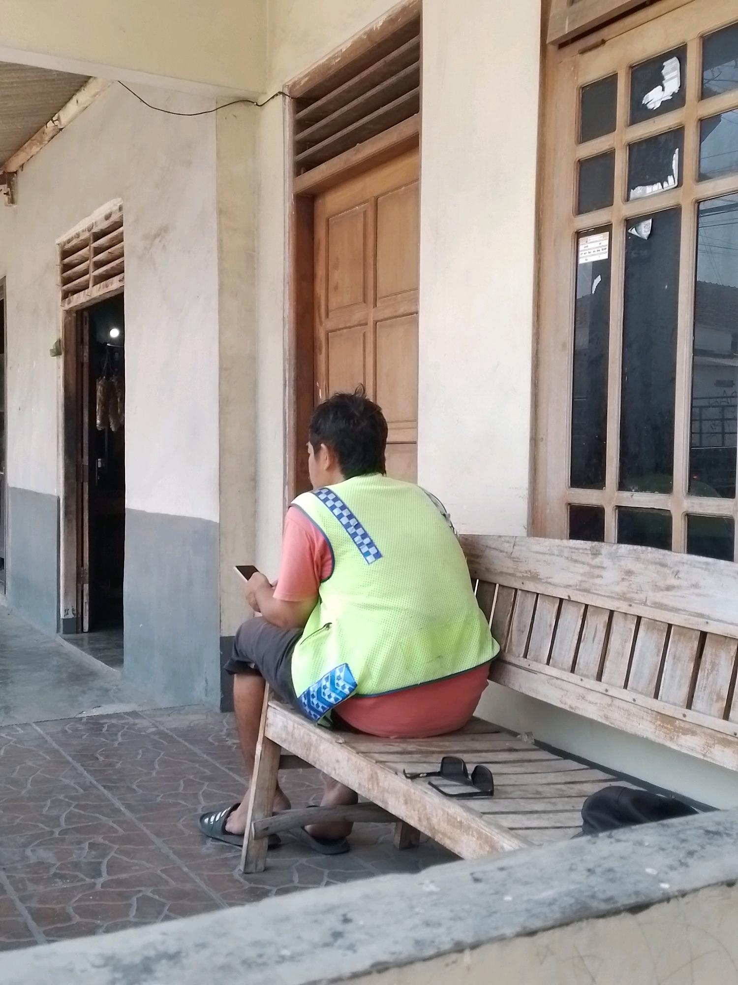 A medium shot captures a person sitting on a wooden bench outside a building in Wonosari, Indonesia. The individual, viewed from the back, is wearing a bright yellow vest over a coral-colored shirt and dark shorts. They are holding a mobile phone in their hands. Sunglasses and a dark cap are placed on the bench next to them. The building features wooden doors and windows with multiple panes, and the ground is tiled with a brown, irregularly patterned design. The lighting suggests it is daytime.