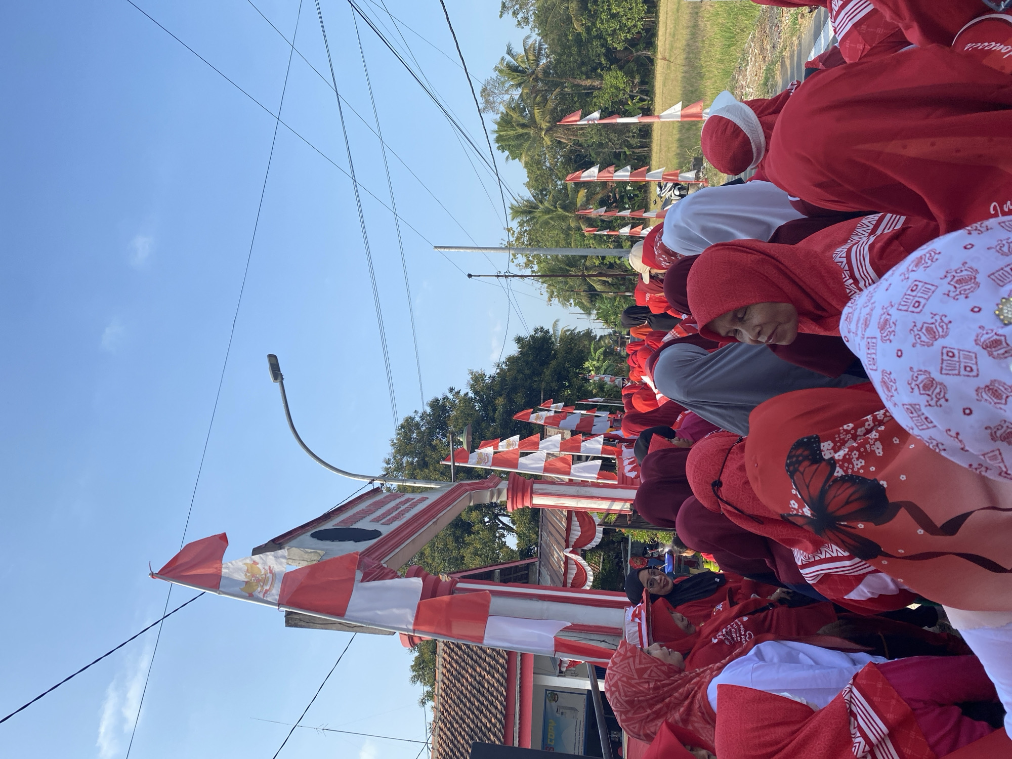 The image depicts a crowd of people, predominantly women, gathered outdoors. Most individuals are wearing red clothing and headscarves, suggesting a coordinated event or celebration. The atmosphere appears festive, with numerous red and white Indonesian flags decorating an archway and lining a path. Trees and a building with a tiled roof are visible in the background, indicating a rural or suburban setting. The sky is clear and blue. The event seems to be taking place in Gumelar Kidul, Indonesia, as suggested by the presence of Indonesian flags and the overall context.