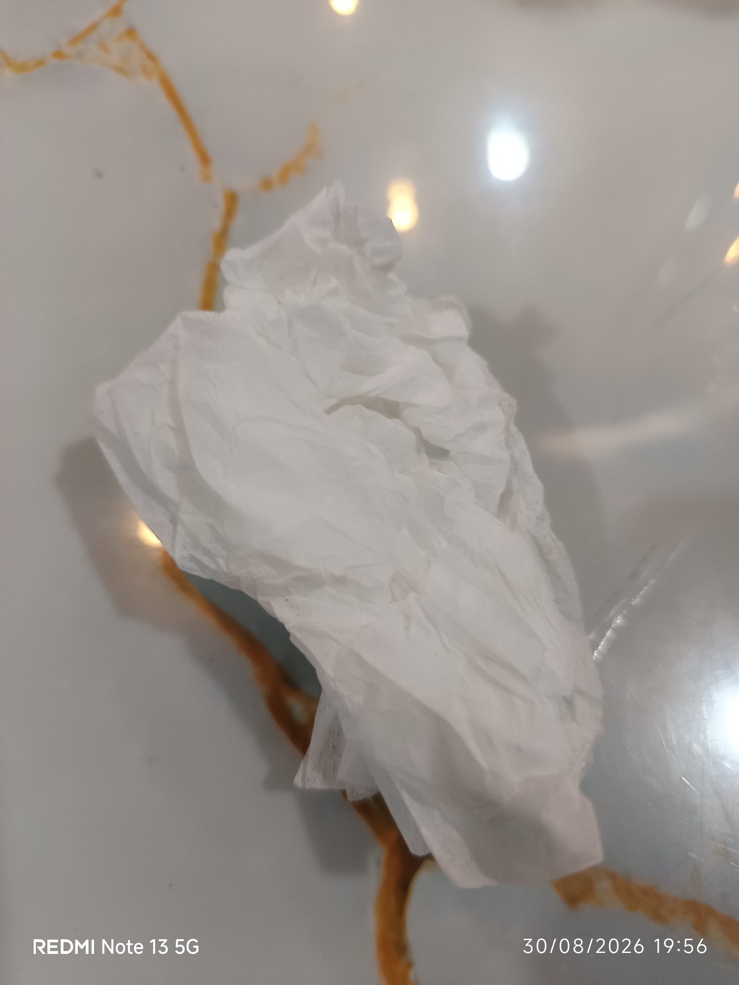 A crumpled white tissue is centrally located on a table with a marble-like surface featuring prominent brown veining. The tissue appears dry and is irregularly shaped. The lighting suggests an indoor environment, with reflections visible on the table surface. The bottom left corner of the image displays the text "REDMI Note 13 5G", and the bottom right corner shows the date and time "30/08/2026 19:56". There are no individuals or animals present in the scene. The location specified, Kota Galuh, Indonesia, is not depicted or inferable from the visual information provided in the image.