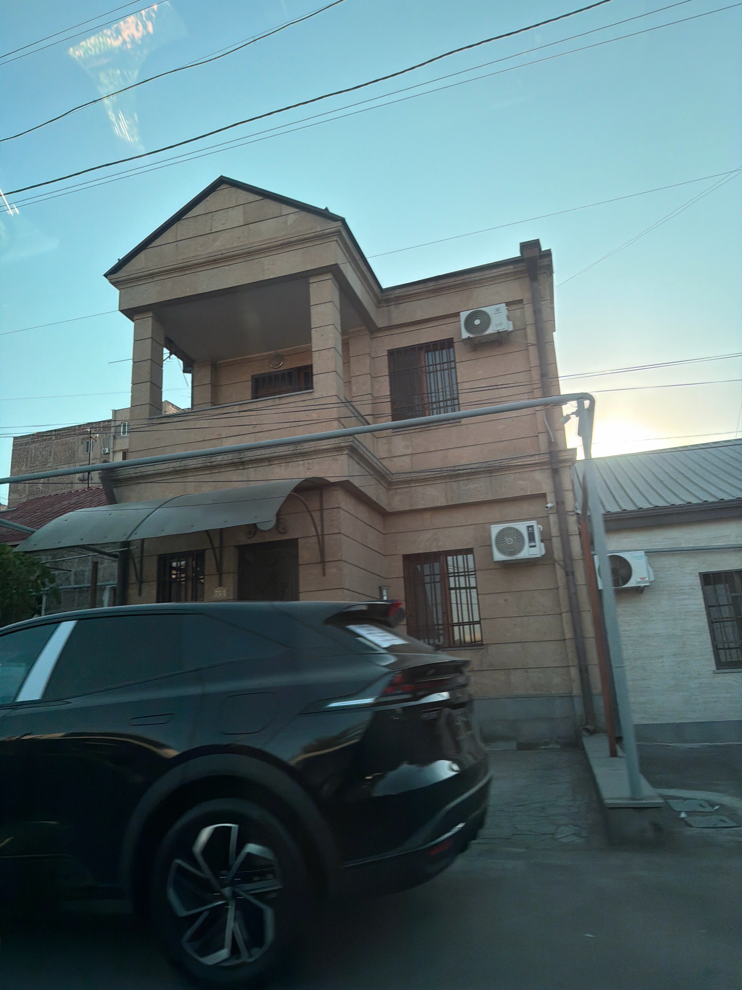 A black SUV is driving on a street in Yerevan, Armenia. The car is in the foreground, and the background shows a building with beige walls and a dark roof. There are also power lines and air conditioners on the building. The sky is blue and clear. The car is moving, and the image is slightly blurred.