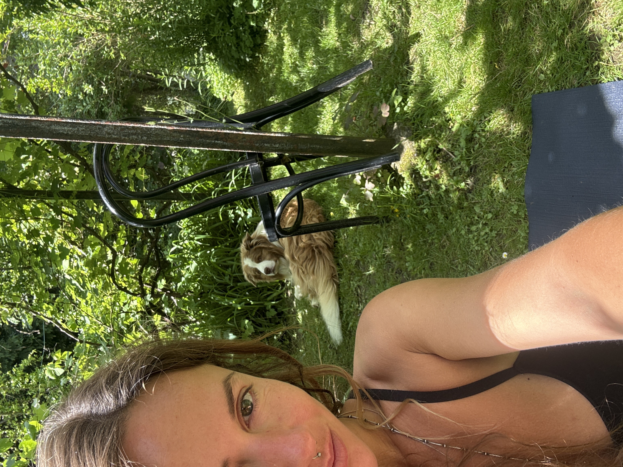 An adult human, identified as a female, is in the foreground, holding a camera in her right hand for a selfie. She has long brown hair, a nose ring, and is wearing a black tank top. Her left arm and shoulder are visible. Behind her, a dog with brown and white fur is lying on a green lawn. The dog is in focus, while the woman is slightly out of focus. In the midground, there is a metal pole and a black metal chair. The environment is a sunny outdoor setting with lush green foliage and grass, suggesting a garden or park. The lighting indicates it is daytime.