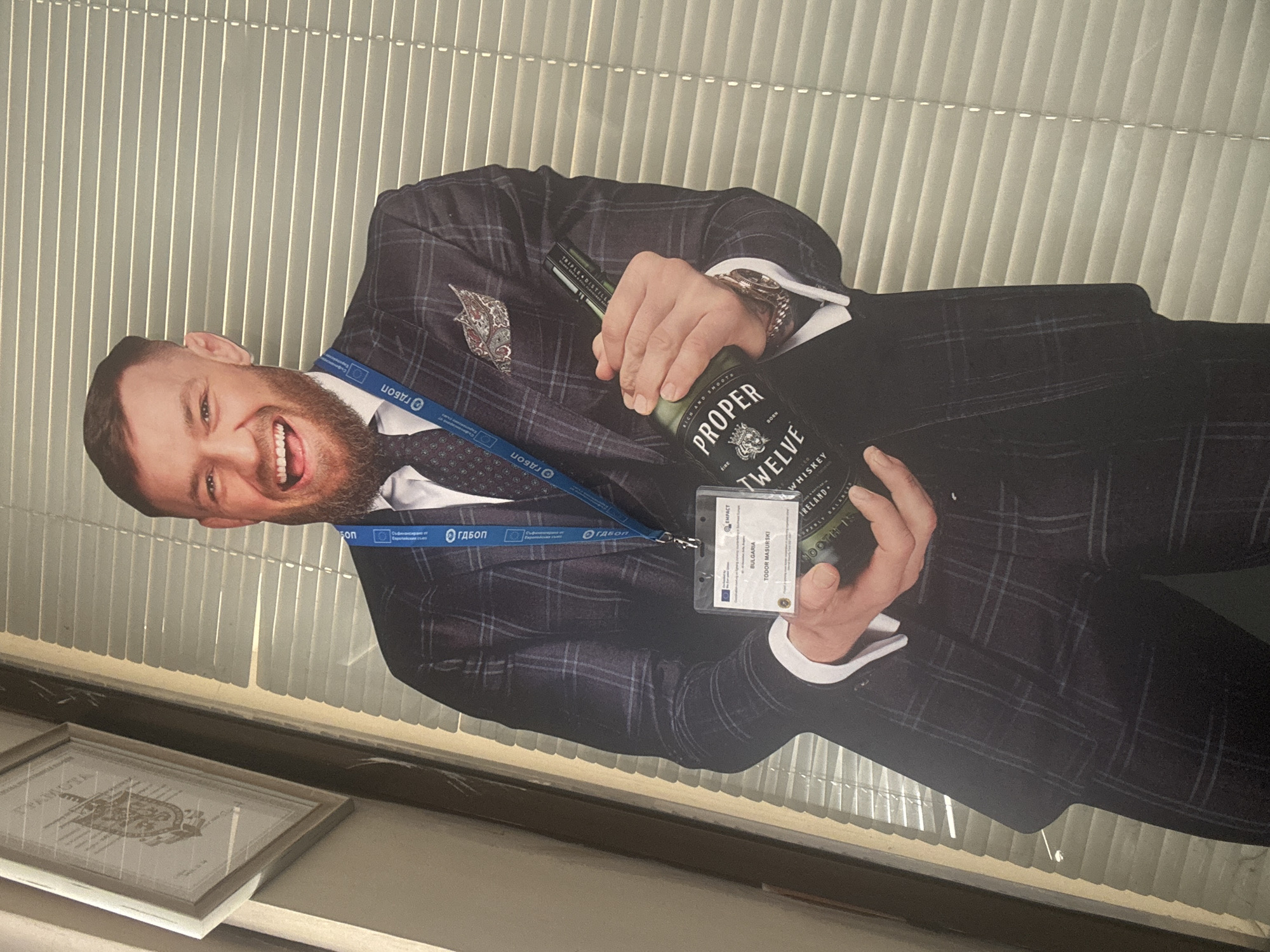 The image displays a promotional poster of Conor McGregor in a suit, holding a bottle of "Proper Twelve" whiskey. He is wearing a lanyard with a name tag that appears to say "Todor Masurski, Bulgaria", along with some logos that include "ГДБОП". The poster is displayed on a ledge in front of blinds. Below the poster is a framed certificate. Given the presence of "ГДБОП" logo on the poster, it's possible the location is related to Bulgarian law enforcement or governmental agencies. Without further information, a specific location in Plovdiv cannot be determined.