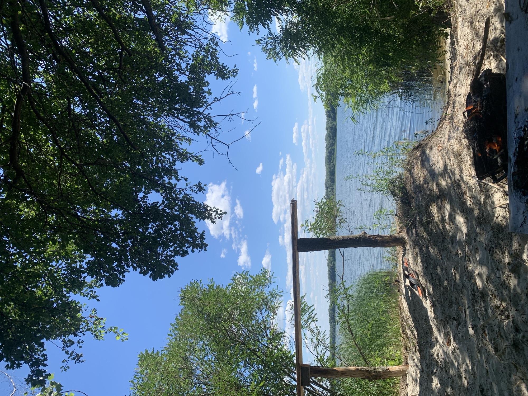 This media file depicts an outdoor scene near a body of water, likely a river or lake, in Horishni Plavni, Ukraine. The image is oriented vertically, suggesting it was taken with a phone camera.
The foreground is dominated by a sandy embankment on the right, with shadows cast by trees. A portable grill with glowing embers and flames is visible on the embankment, indicating a barbecue or campfire is in progress. To the left of the grill, a pair of orange and black flip-flops are resting on the sand. Further left and in the foreground, the branches and foliage of leafy trees frame the scene.
The middle ground features the water, which appears calm with gentle ripples. Lush green vegetation, including tall reeds or grasses, lines the edge of the water. A rustic wooden structure, possibly a makeshift bench or part of a dock, is also present near the water's edge.
The background shows a distant tree line across the water and a bright blue sky dotted with fluffy white clouds. The lighting suggests it is daytime, likely late morning or early afternoon, under fair weather conditions. The overall impression is one of a relaxed, outdoor leisure activity by the water. There is no visible text in the image.