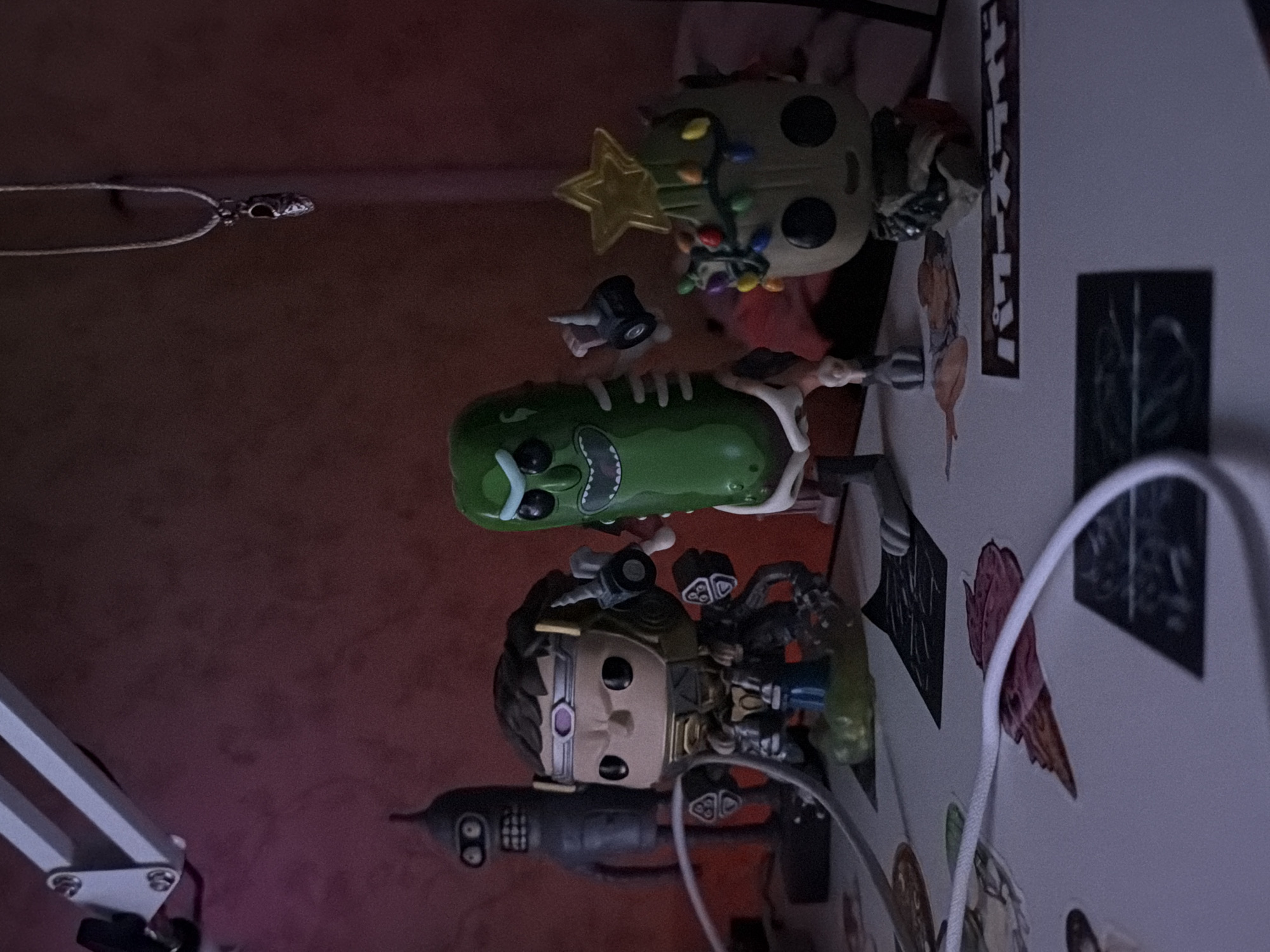 The image displays a collection of pop culture figurines arranged on a surface. From left to right, the figurines include a Bender from Futurama, a character with brown hair and a purple forehead jewel, a green pickle character with skeletal features, and a Groot figurine decorated with Christmas lights and a star. A white cable snakes across the surface in front of the figurines. In the background, a desk lamp with a white arm is visible, casting a shadow on a reddish-pink textured wall. A thin silver necklace with a pendant hangs from the upper right of the frame. The scene appears to be set indoors on a desk or shelf, in an environment that is dimly lit with red and white light sources.