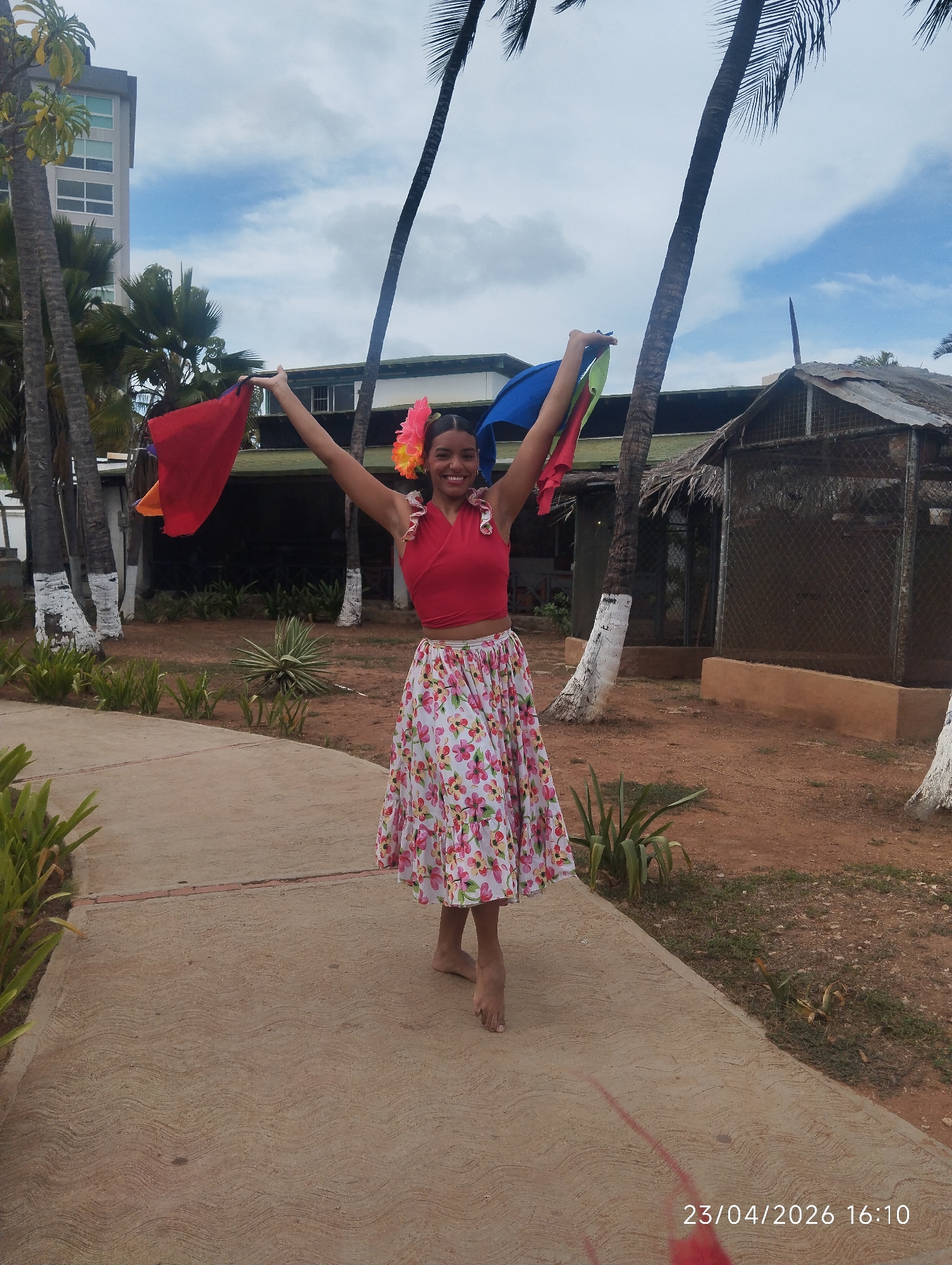 The image displays a vibrant outdoor scene in what appears to be a tropical setting, consistent with Porlamar, Venezuela. The central subject is a young woman standing on a light-colored concrete path with a distinctive wavy texture. She is smiling broadly and joyfully, with her arms raised, holding up a red piece of fabric in her left hand and a blue one in her right.
She is dressed in a bright red crop top and a white, flowing skirt adorned with a colorful floral pattern featuring pink, green, and yellow blooms. Large pink and yellow flowers are tucked into her hair on the right side of her head. She is barefoot.
The background features several tall palm trees, some with their bases painted white, and various tropical plants growing in reddish-brown dirt. In the upper left, a multi-story modern building with numerous windows is visible beneath a partly cloudy sky. To the right, a rustic structure with a thatched roof and wire mesh enclosures can be seen.
The activity suggests a moment of celebration, dance, or performance, indicated by the woman's pose, attire, and the colorful fabrics she holds. The time of day is late afternoon, as confirmed by the timestamp "23/04/2026 16:10" in the bottom right corner. The sky shows a mix of blue and white clouds, indicating partly cloudy weather. The overall impression is one of happiness and cultural vitality in a warm, outdoor environment.