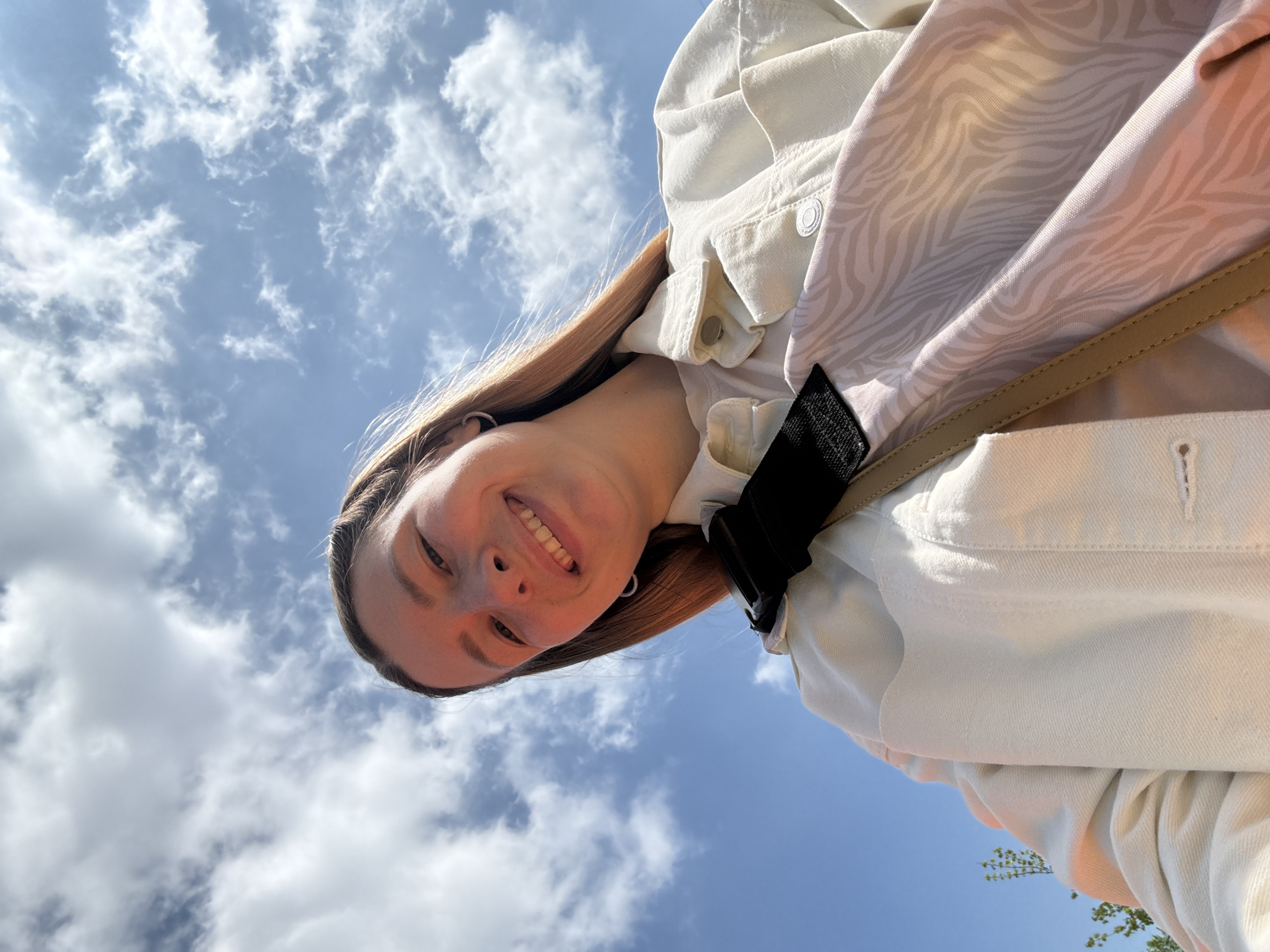 A low-angle shot captures a smiling young woman against a backdrop of a blue sky with scattered white clouds. She has fair skin and long, straight brown hair. She's wearing a light-colored jacket with a pale zebra-print lining, and a tan-colored strap is visible across her shoulder. The background is dominated by the sky, hinting at an outdoor location. Moscow, Russia is not explicitly evident in the image.