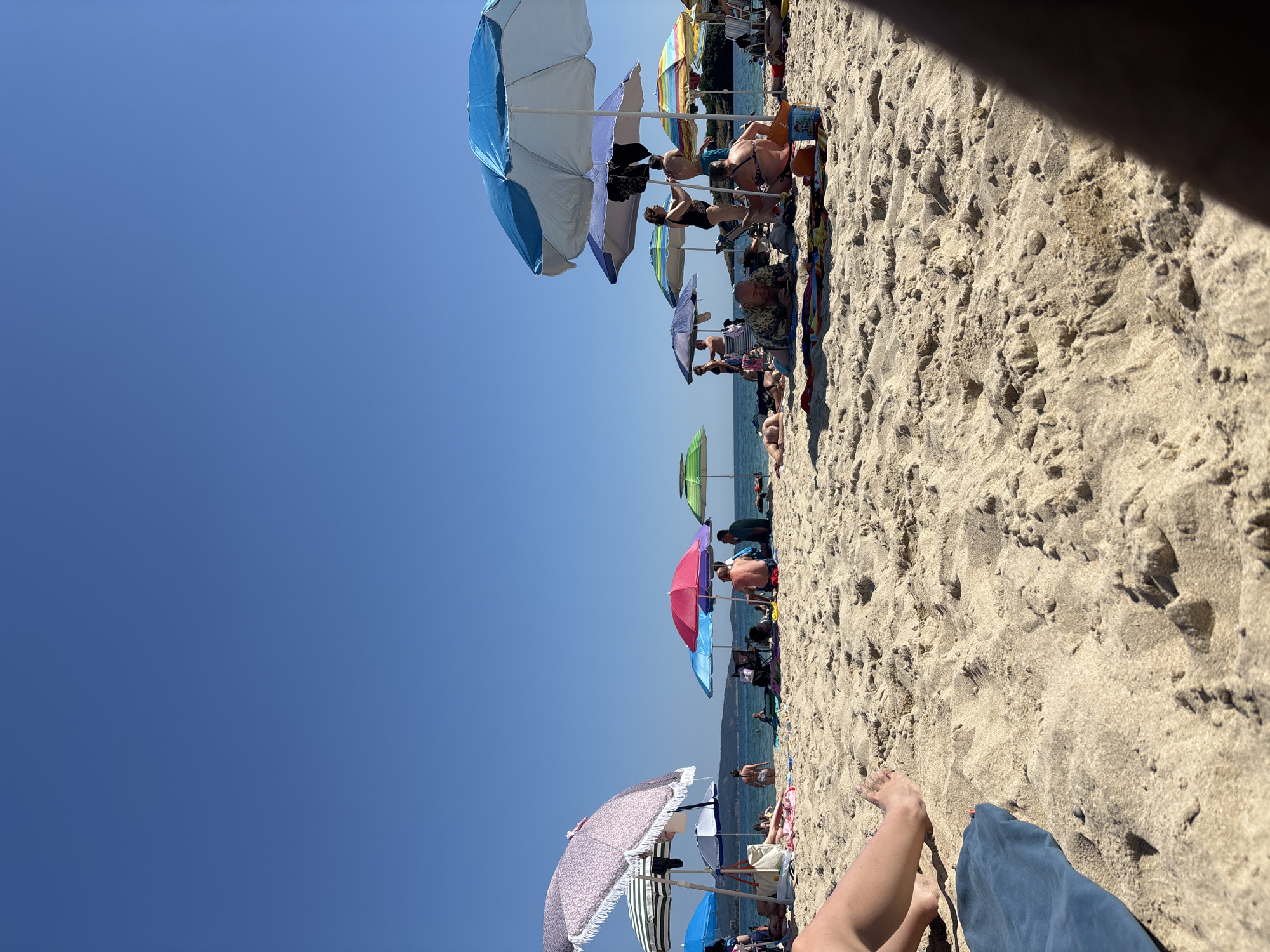 The image depicts a sunny day at a crowded beach in Kiten, Bulgaria. The foreground is dominated by coarse, light-colored sand, with a blue towel visible on the left. A person's legs and feet are also present in the lower left corner. In the middle ground, numerous beachgoers are spread across the sand, many seated or lying under various colorful umbrellas. The umbrellas are a mix of styles and colors, including bright blue, pink, green, and a patterned one with fringes. Some people are in the water, which is visible in the background, appearing calm and blue under a clear, bright sky. The overall atmosphere suggests a typical summer day at a popular tourist destination.