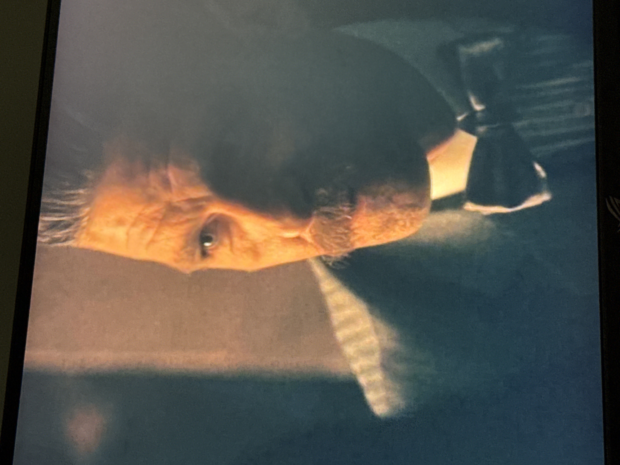 A close-up, rotated image shows a man's face. The man is elderly with wrinkles and fair skin. He has gray hair. He is wearing a dark suit and tie with a white shirt collar visible. The background is blurred and dark. The location is Unknown, Unknown.