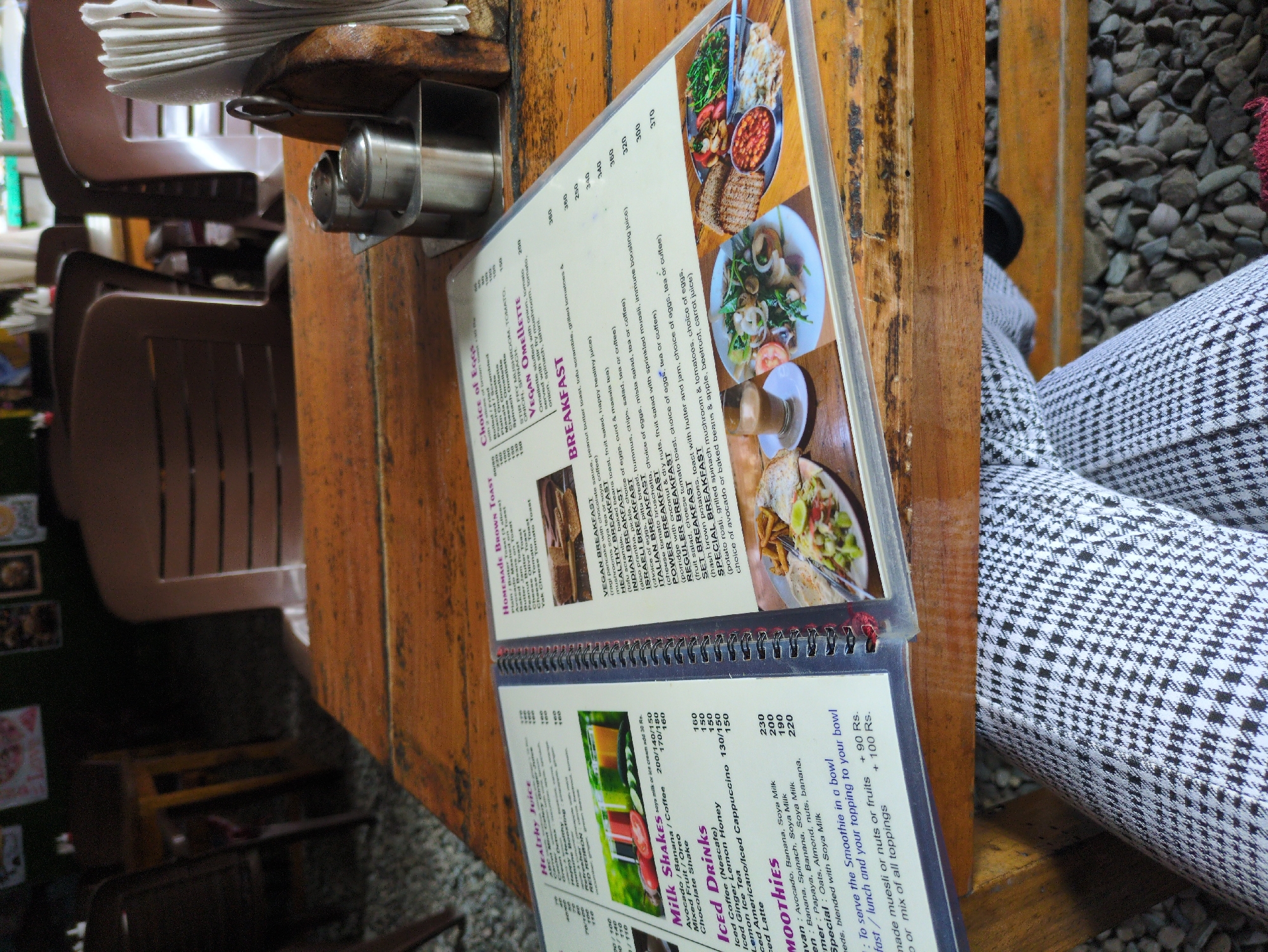The image shows a table in a cafe in Bhagsu, India, with a menu open to the breakfast and drinks sections. The table is made of rough wood. On the table, there is a napkin holder with white napkins, and two metal salt and pepper shakers. The menu has images and text. The drinks section lists "Healthy Juice," "Milk Shakes," "Iced Drinks," and "Smoothies," with prices. The breakfast section lists "Homemade Brown Toast," "Choice of Eggs," "Vegan Omelette," and various "Breakfast" options with descriptions and prices. The background shows chairs and a glimpse of the cafe's interior, possibly with other patrons, though no people are clearly visible in the foreground. The overall impression is a casual dining setting.