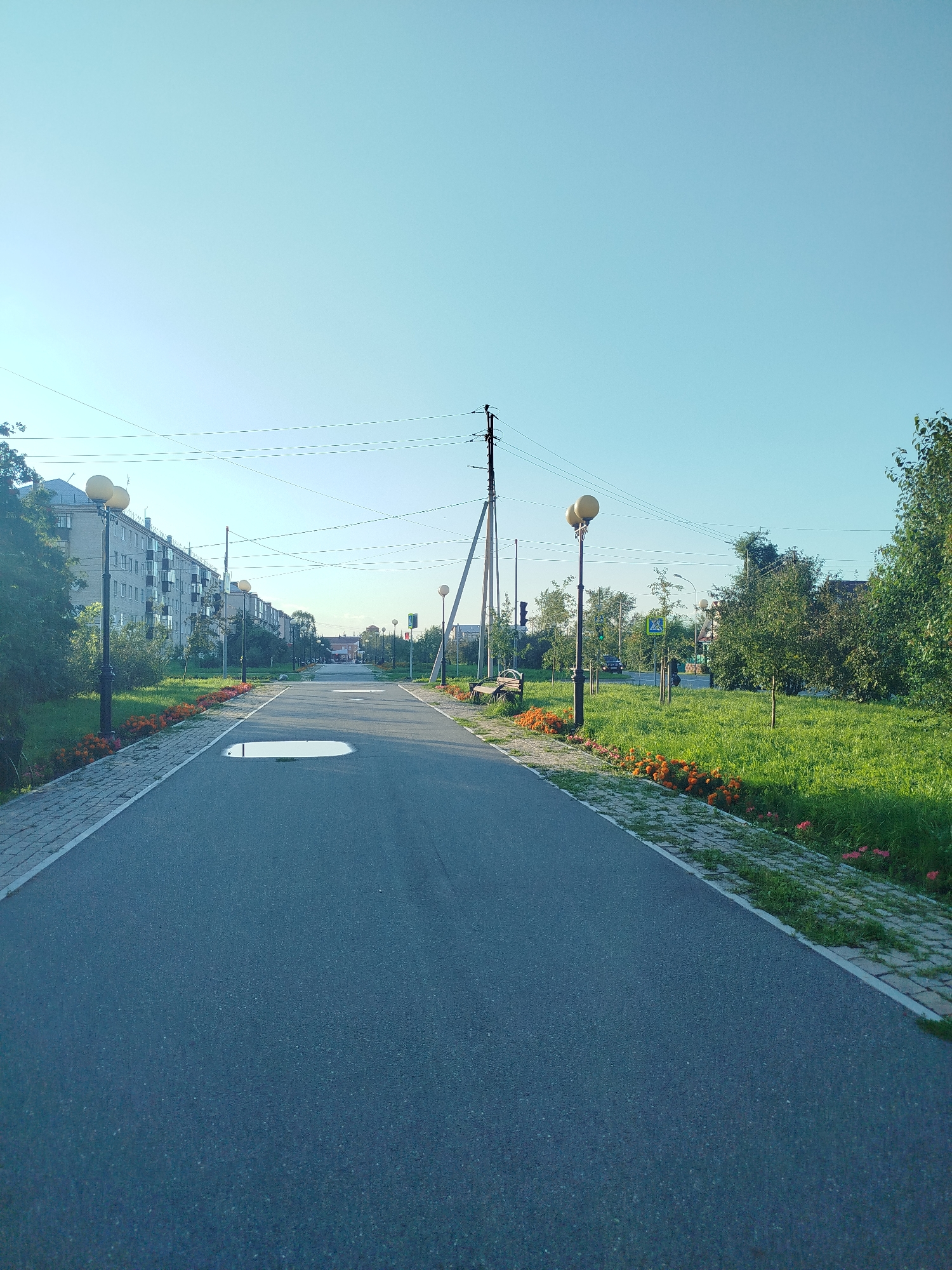 The image depicts a wide asphalt pathway in a city park, likely in Yalutorovsk, Russia, on a clear day. The pathway is lined with green grass, flowerbeds containing orange flowers, and interspersed with trees and streetlights. In the background, there are multi-story apartment buildings and a street with some vehicular and pedestrian activity. The sky is a clear, pale blue, indicating a pleasant weather condition. The pathway itself has a large puddle of water on it, suggesting recent rain or watering. There are no people clearly visible on the pathway or in the immediate surroundings. The scene is characterized by the presence of urban infrastructure within a green, outdoor environment.