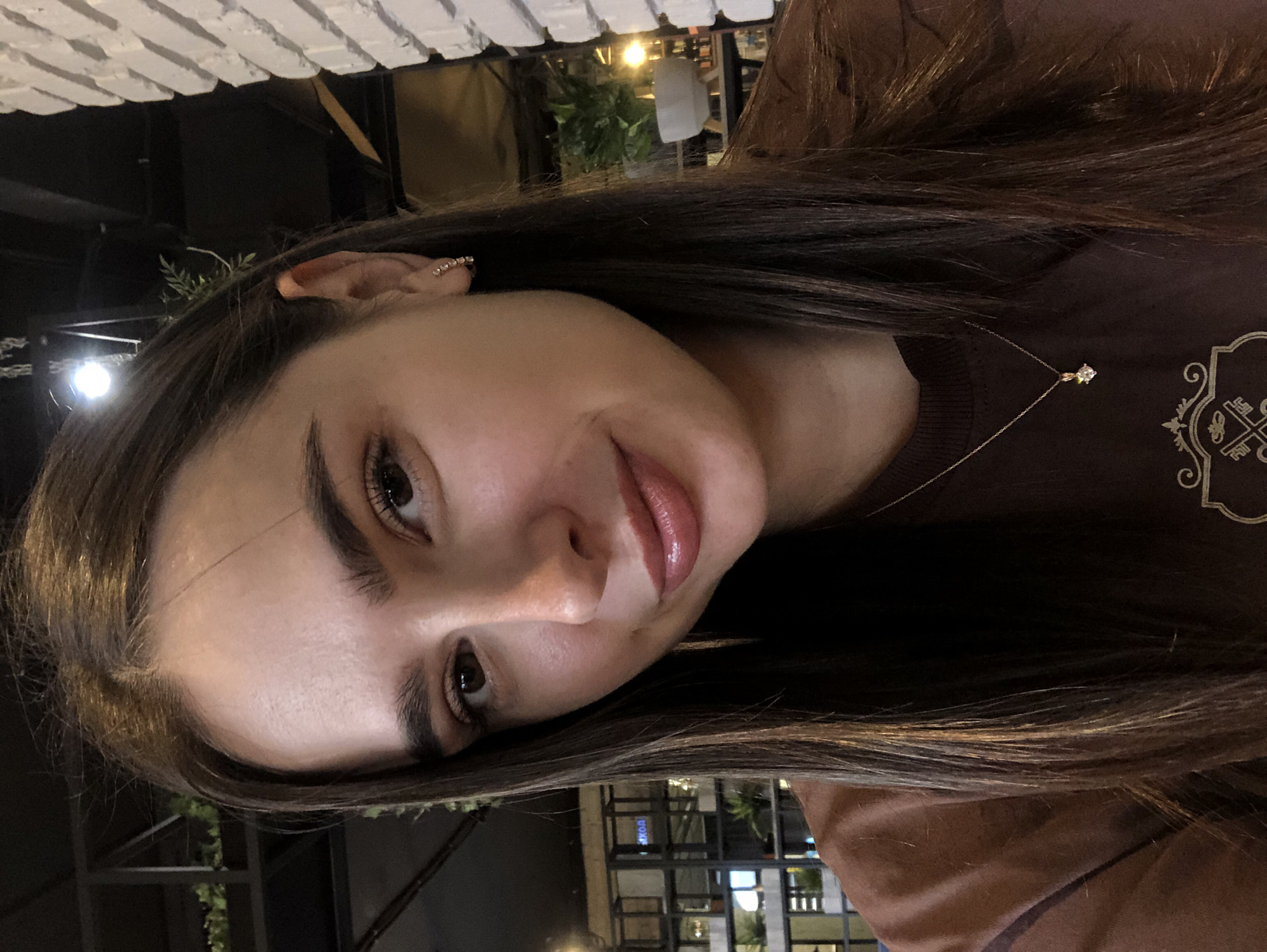A young woman with long, dark brown hair, wearing a brown sweatshirt and a gold necklace with a small diamond pendant, smiles at the camera. She has on subtle makeup, including mascara, blush, and a glossy pink lipstick. Her right earlobe is adorned with a small hoop earring. The background suggests an indoor setting, possibly a cafe or restaurant, with exposed brick walls on the right and a dark ceiling with lighting fixtures and hanging plants. To the left, there are shelves with plants and glass doors, hinting at a commercial or public space. The image appears to be a selfie, taken from a close-up perspective, focusing on the woman's face and upper torso. There is no indication of the specific location within Izhevsk, Russia.