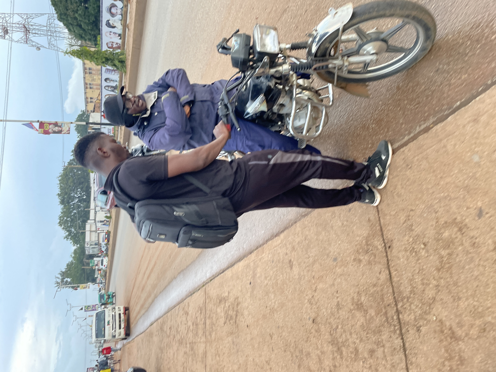 Two men are standing by a motorcycle on a dirt and asphalt road in Jos, Nigeria. The man on the left is wearing a black t-shirt, black track pants with a gray stripe, and black sneakers. He has a black backpack on his shoulders. The man on the right is wearing a blue jacket and pants, a black baseball cap, and glasses. He has his arms crossed. The motorcycle is black and silver with chrome accents. The road is lined with a sidewalk made of reddish-brown dirt. In the background, there are buildings, trees, streetlights, a cellular tower, and a banner hanging from a pole. Vehicles, including a white van and a green tricycle, are visible on the road. The sky is overcast.