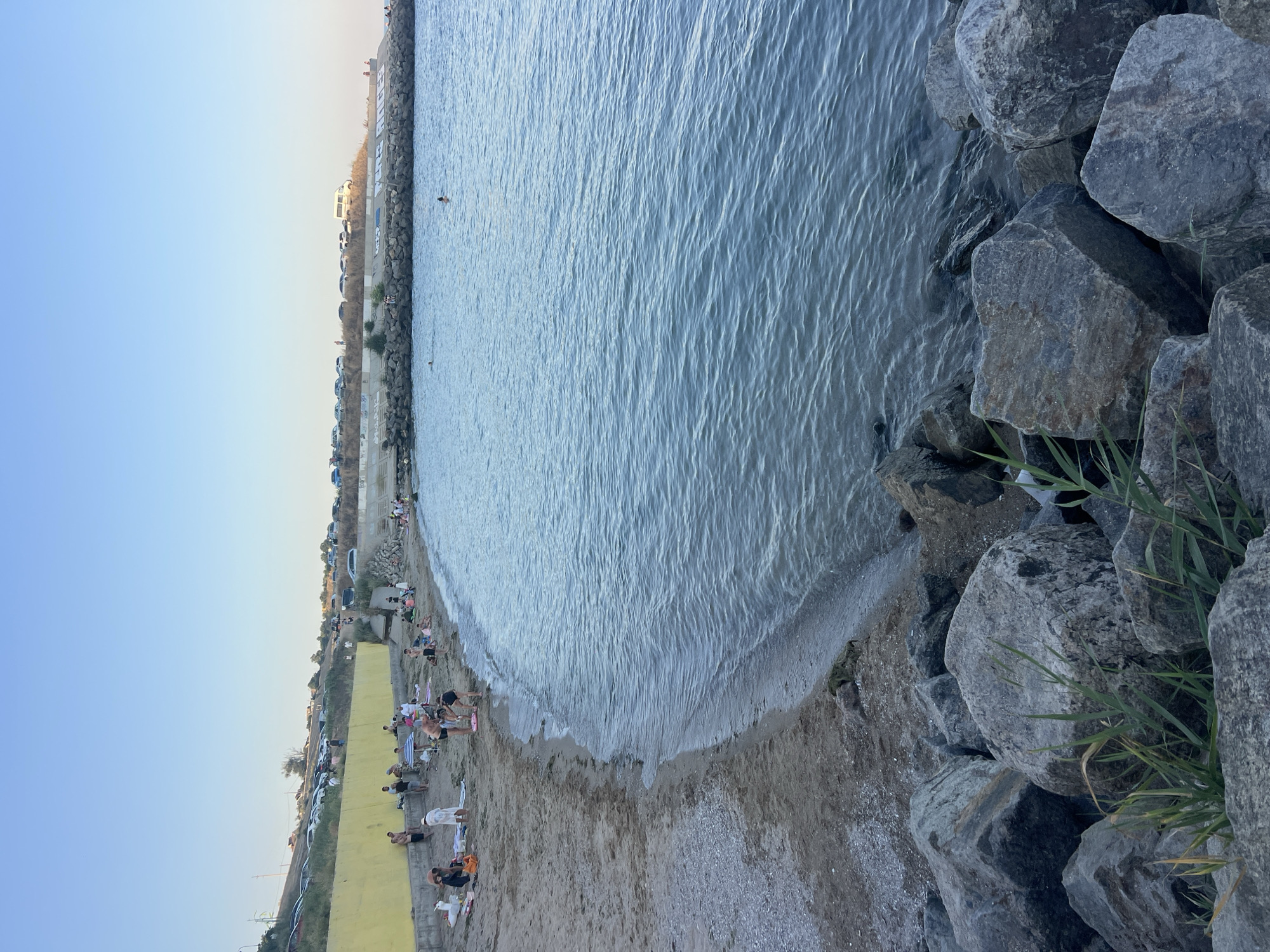 A group of people are on a sandy beach near a body of water. The beach has a yellow wall along its length, with a parking lot and a road visible behind it. The water is calm with some small waves. In the foreground, there are large rocks. Some people are sitting on towels, while others are standing or walking. Two individuals are in the water. The sky is clear and blue, suggesting it is daytime.