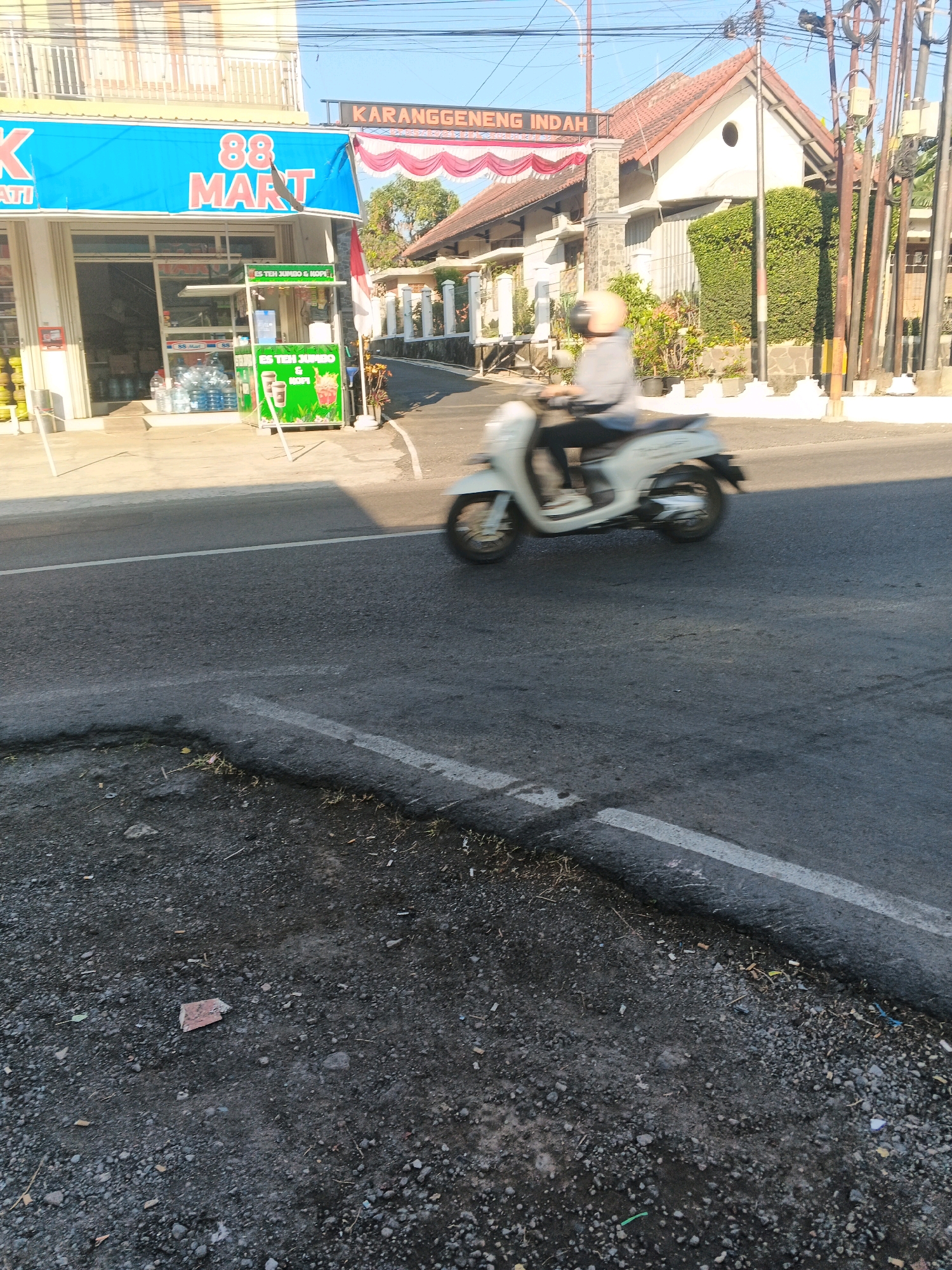 The image shows a street scene in Semarang, Indonesia, with a storefront on the left and a residential building on the right. In the foreground, a white scooter with a rider is moving across the asphalt road, which shows signs of wear and tear with cracks and loose gravel along the edges. The rider, wearing a light-colored shirt and a helmet, is blurred due to motion.
The storefront on the left is marked with a large blue sign that reads "88 MART". Below the sign, the store entrance is visible, with shelves stocked with goods, including bottles of water. A green kiosk with images of drinks is positioned outside the store.
The building on the right has a reddish-brown tiled roof and a white facade. A sign above the entrance of this building reads "KARANGGENENG INDAH" in orange lettering. The area around the building is landscaped with greenery and potted plants. Overhead, power lines crisscross the sky, and a street lamp is visible on the right side of the image. The overall lighting suggests it is daytime, with the sun casting shadows on the road.