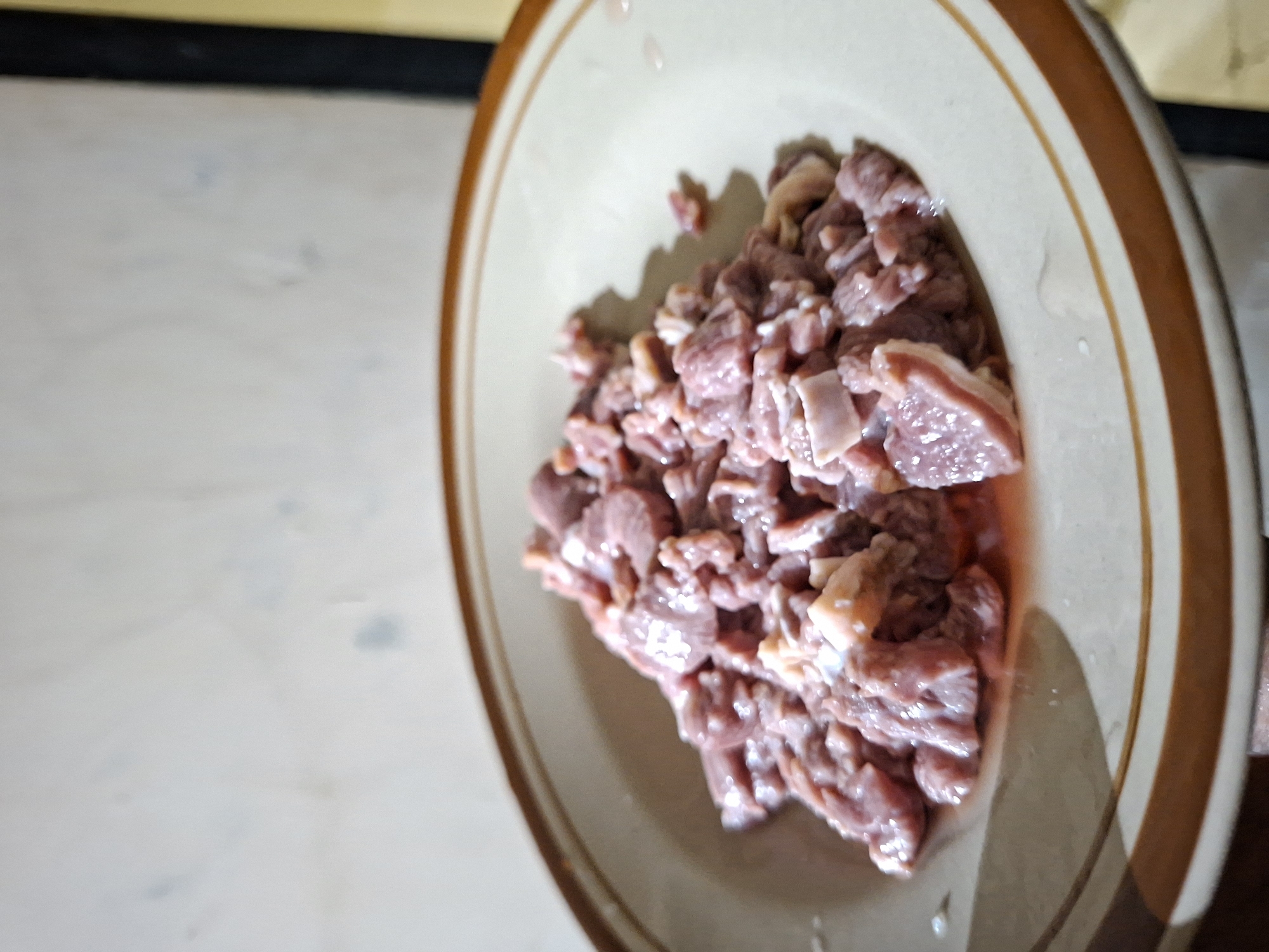 The image shows a close-up view of a plate filled with raw, diced meat. The plate is cream-colored with a brown rim. The meat appears to be a mixture of lean muscle and some fat. There is a small amount of liquid present in the plate with the meat. The background is a light-colored surface. There are no people visible in the image. It is not possible to confirm that the image was taken in Lhokseumawe, Indonesia.