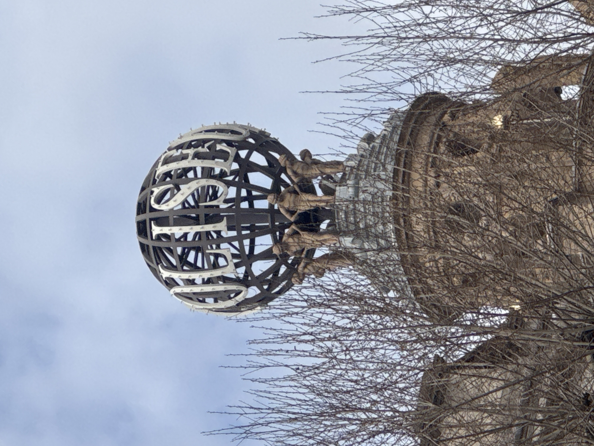 A low-angle shot reveals a complex sculpture situated near a stone structure, likely part of a building, in what appears to be London, United Kingdom. The primary sculpture is a spherical cage-like structure with intertwined words or designs. Several small figures are positioned around the circumference where the sphere connects to the building. Bare branches of trees are visible in the foreground, partially obscuring the view of both the sculpture and the building. The sky in the background is overcast.
