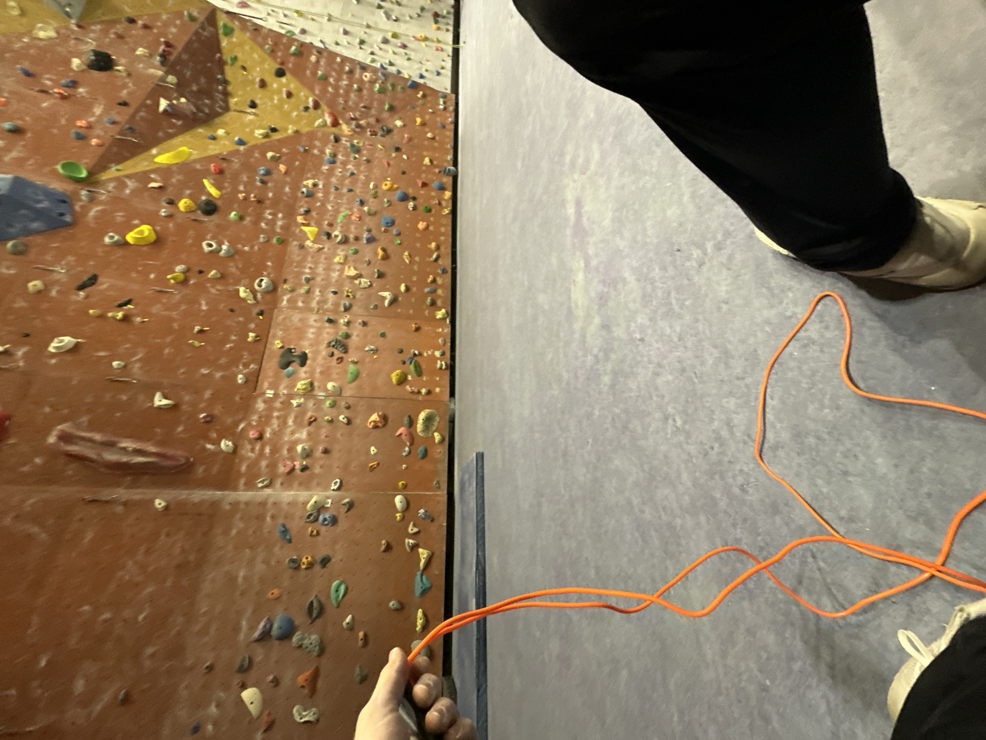 The image shows a person's perspective inside what appears to be a rock climbing gym. On the left is a climbing wall covered in holds of various sizes, shapes, and colors. The wall itself is orange and appears textured. A portion of the climber’s hand is visible at the bottom of the wall, holding a thin, orange cord.
On the right, the image shows the gym floor, which is light grey and textured. A person’s legs, clad in black pants and tan shoes, are standing on the floor. An orange cord is snaked across the floor near their feet.
Without additional context, it is not possible to confirm the location within the city of Sofia, Bulgaria.