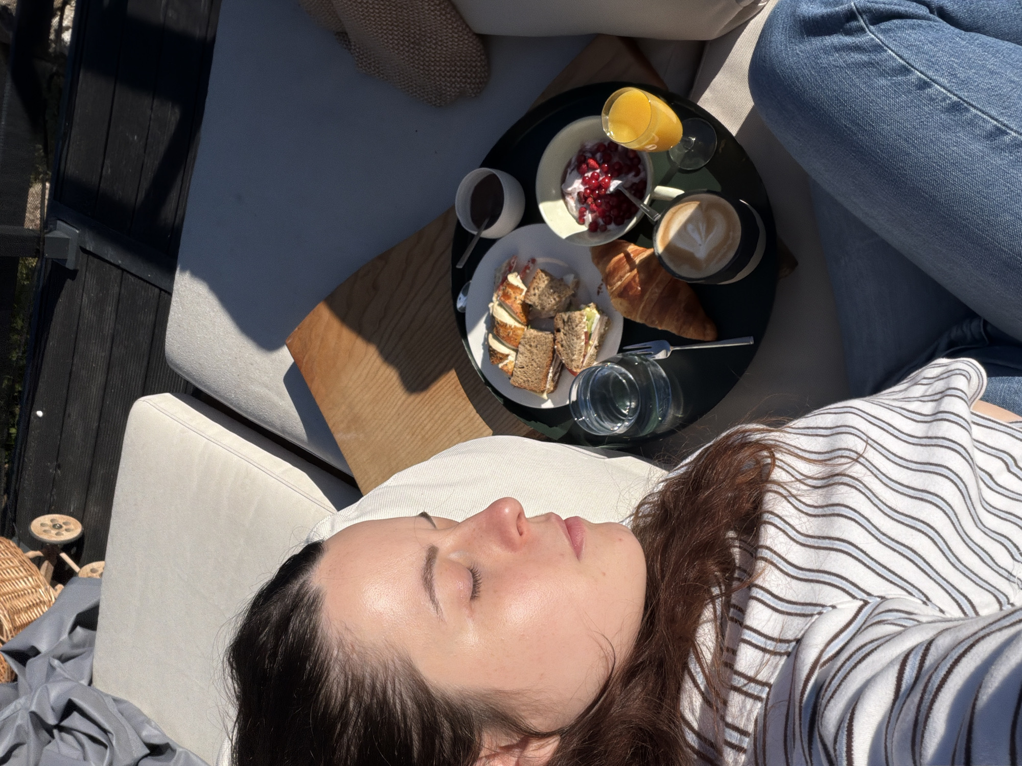 A woman is lying on an outdoor lounger, eyes closed, with a breakfast tray placed on her lap. The tray contains a croissant, a bowl of yogurt with berries, a cup of coffee with latte art, a glass of orange juice, a glass of water, a fork, and a small dish of chocolate sauce. There is also a plate with what appears to be a sandwich and some sliced bread. The background suggests an outdoor setting, possibly a deck or patio, with wooden planks and a dark railing. The sunlight indicates it is daytime. The location is not explicitly identified as Gangloff, Germany.