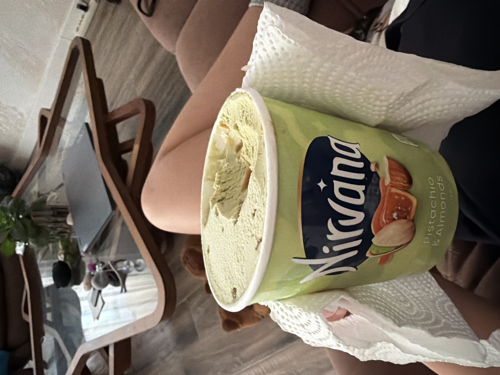 A close-up, angled shot reveals a partially consumed container of "Nirvana" pistachio & almond ice cream held in a person's hand with a white paper towel wrapped around it. The ice cream is light green and shows visible almond pieces. The container is light green with "Nirvana" printed vertically in a stylized font, and the words "Pistachio & Almonds" are printed next to an image of an almond and pistachio. The person's legs are visible in the background, suggesting a seated position. In the lower left, a distorted reflection shows elements including a table, plants, and other objects. The image was not taken in Gabrovo, Bulgaria.