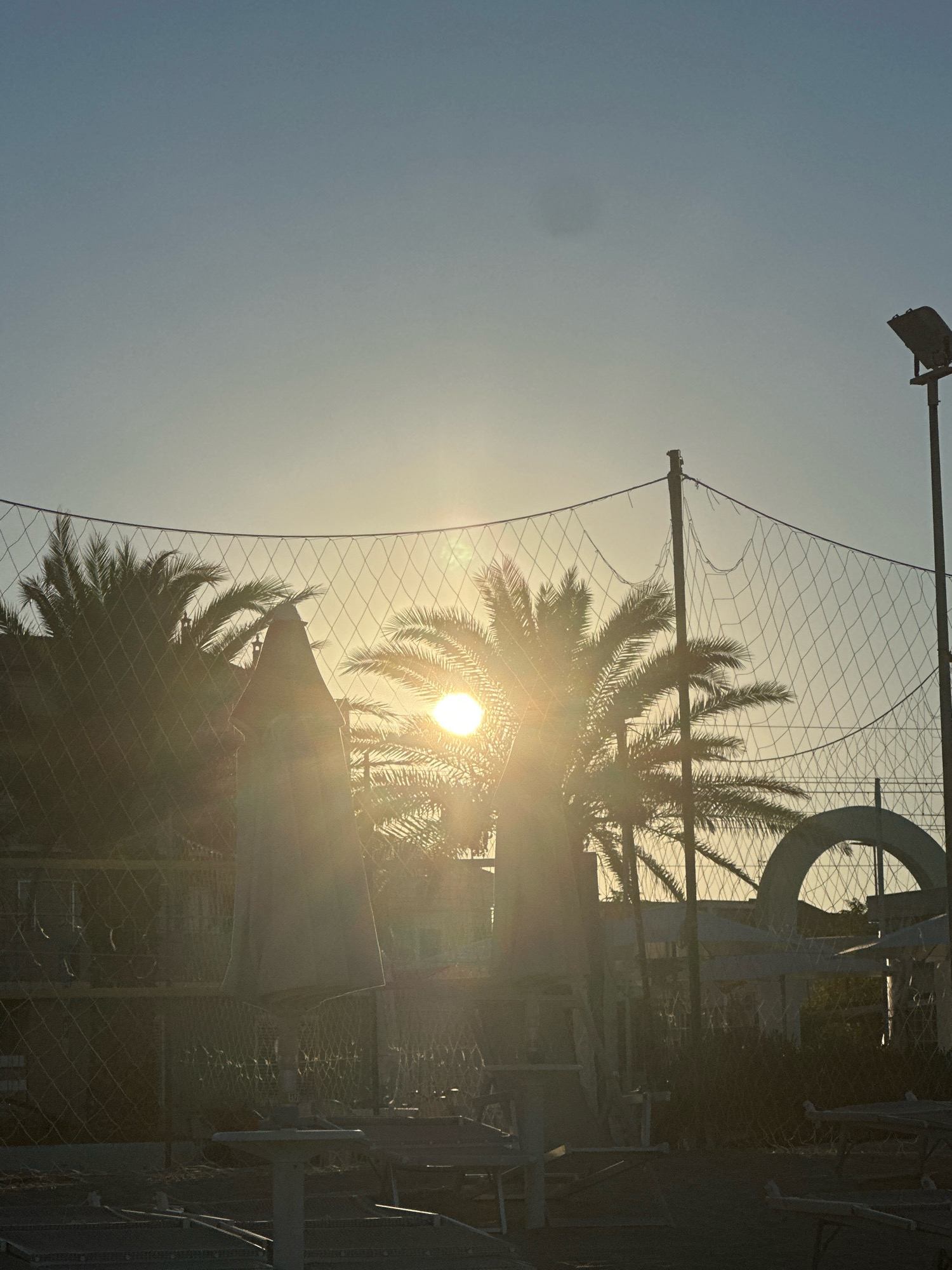 The image depicts a beach scene in Civitanova Marche, Italy, during sunset. The sun is low in the sky, casting a warm glow and lens flare through palm trees and a volleyball net. In the foreground, beach chairs and umbrellas are visible, some appear to be disassembled or in disarray. In the background, a building and an arched structure can be seen. The overall atmosphere is peaceful and serene, with the natural beauty of the sunset dominating the scene.