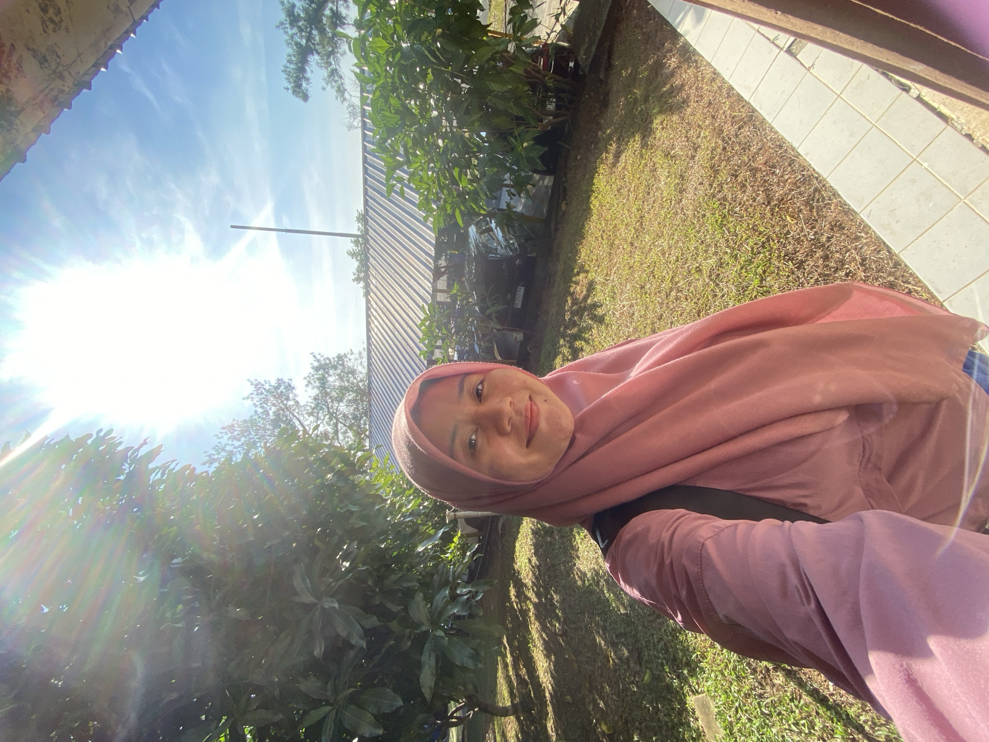 In Banjarbaru, Indonesia, a young woman wearing a pink hijab and a pink top stands for a selfie outdoors on a sunny day. The woman's face is visible and she is smiling. She has a black backpack on. The background shows a lawn with scattered trees and a building with a corrugated metal roof. The sky is blue with some clouds, and the sun is shining brightly, causing lens flare. The scene appears to be in a residential or semi-rural area.