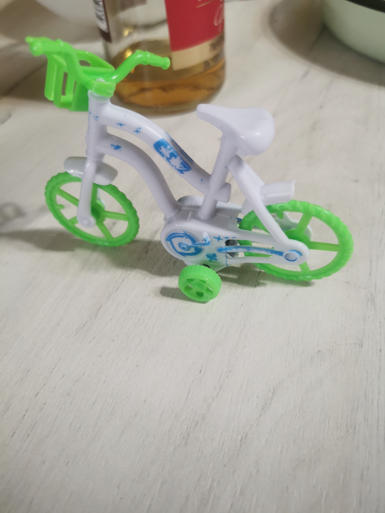 On a white wood-grained surface, a toy bicycle is visible. It is predominantly white with green wheels, handlebars, and training wheels. The toy is positioned at an angle, facing slightly to the left. Traces of a glass bottle are visible in the background, along with a small portion of what appears to be a light green bowl. The location is unknown, and the description provided is independent of any specific location in Stara Zagora, Bulgaria.