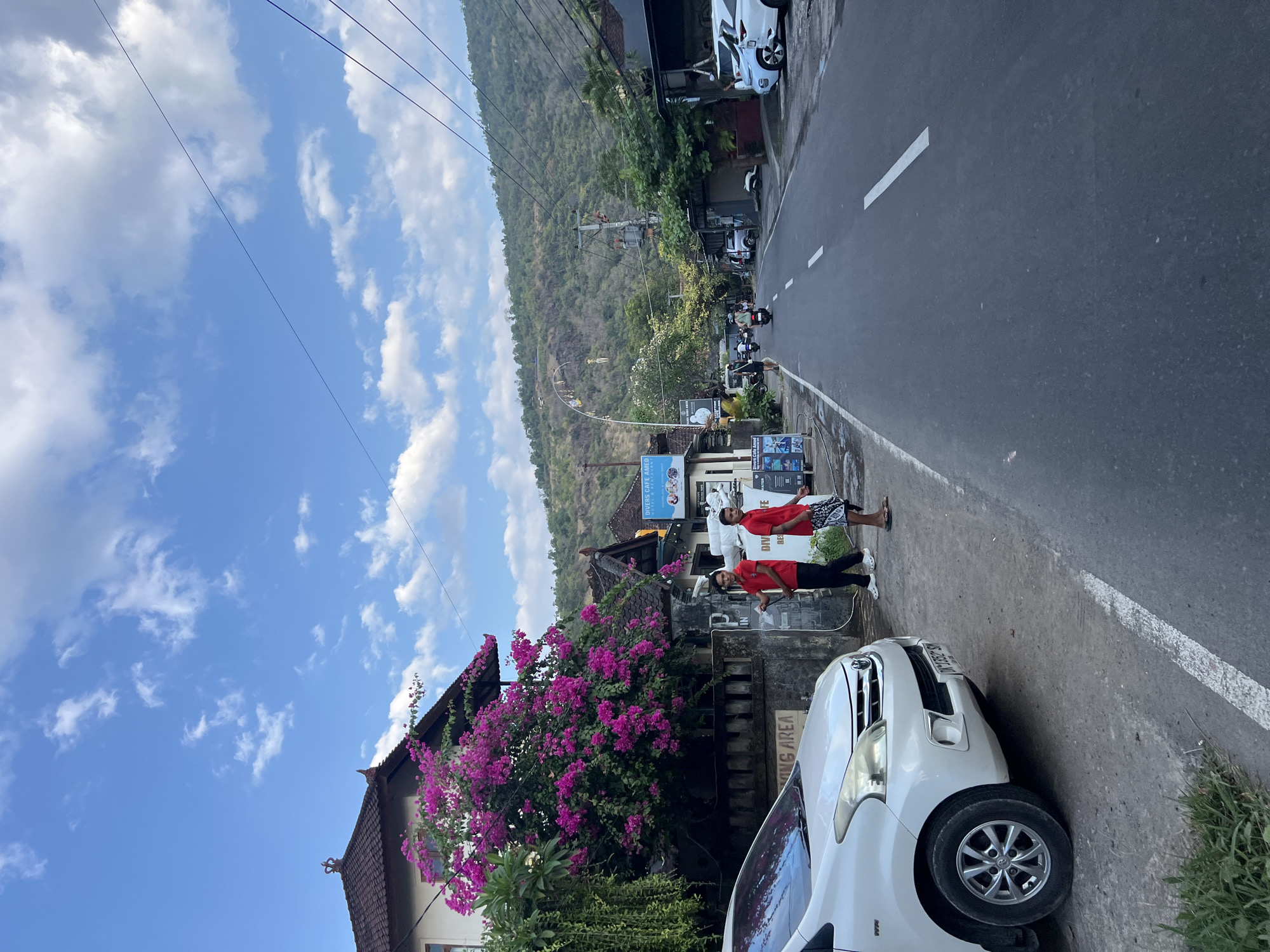 This image depicts a street scene in Abang, Indonesia. A white car is parked on the left side of the road. Two men in red shirts are standing on the sidewalk near a building adorned with bright pink bougainvillea flowers. The road stretches into the distance, with several buildings and trees lining its sides. In the background, a lush green hillside rises under a bright blue sky with scattered clouds. Several motorcycles are visible on the road, suggesting ongoing traffic. Signs are present on some of the buildings, indicating the presence of businesses. The overall impression is one of a quiet, sunny day in a tropical locale.