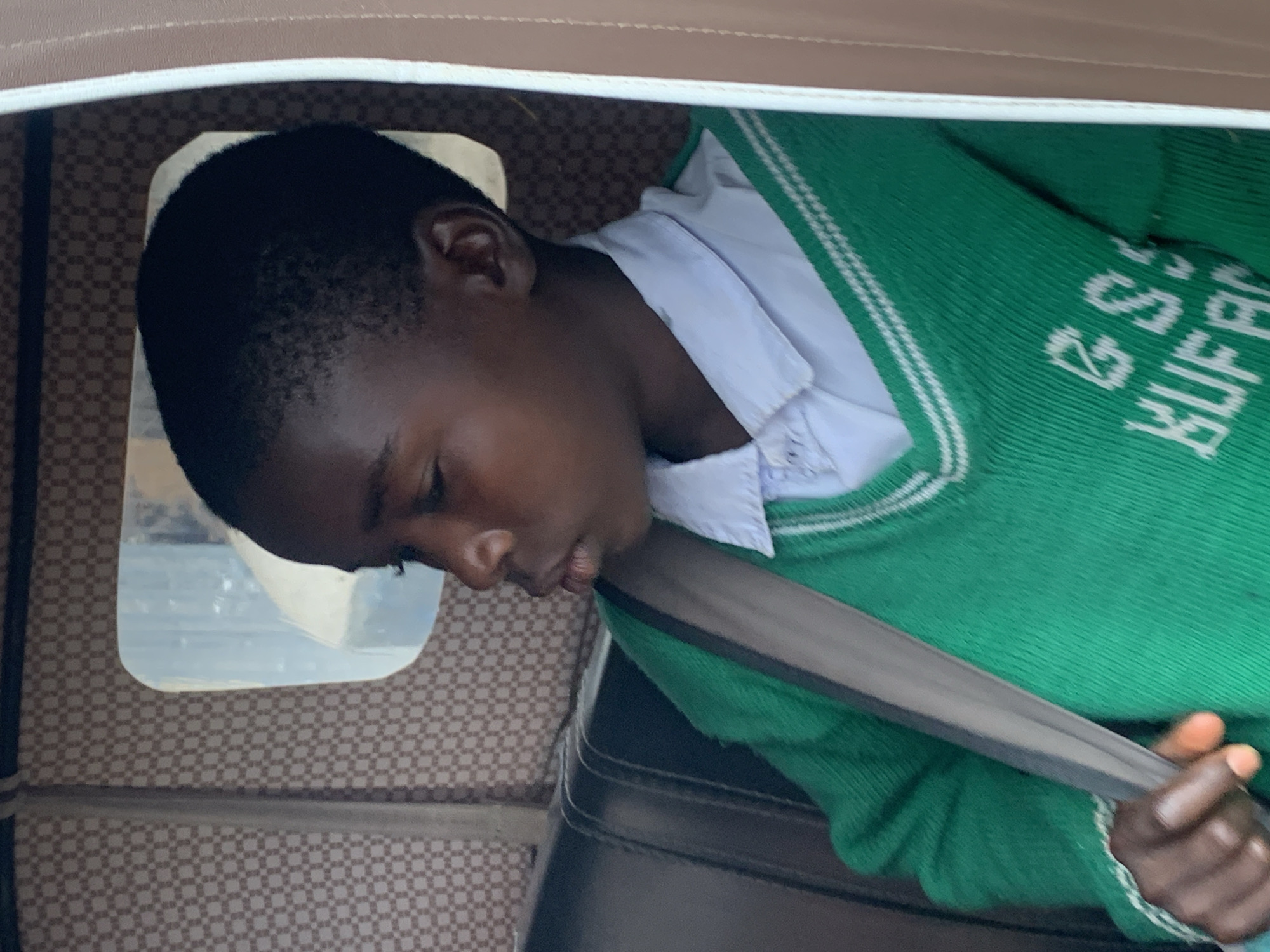 A young boy, appearing to be of school age, is seated inside a vehicle, likely a tuk-tuk given the interior design and visible window. He is wearing a white collared shirt underneath a green V-neck sweater. The sweater has white striped trim on the V-neck and sleeves, and embroidered white text on the front which appears to say "G.S.S BUFA". He is looking to his left, with his right hand resting on his lap, visible as he clutches a grey strap that goes across his chest, possibly a seatbelt or part of a bag. The interior of the vehicle is decorated with a patterned fabric lining the upper walls and a black seat. A window behind the boy reflects a bright, outdoor scene, suggesting it is daytime. The scene is likely captured in Jos, Nigeria, given the context provided.
