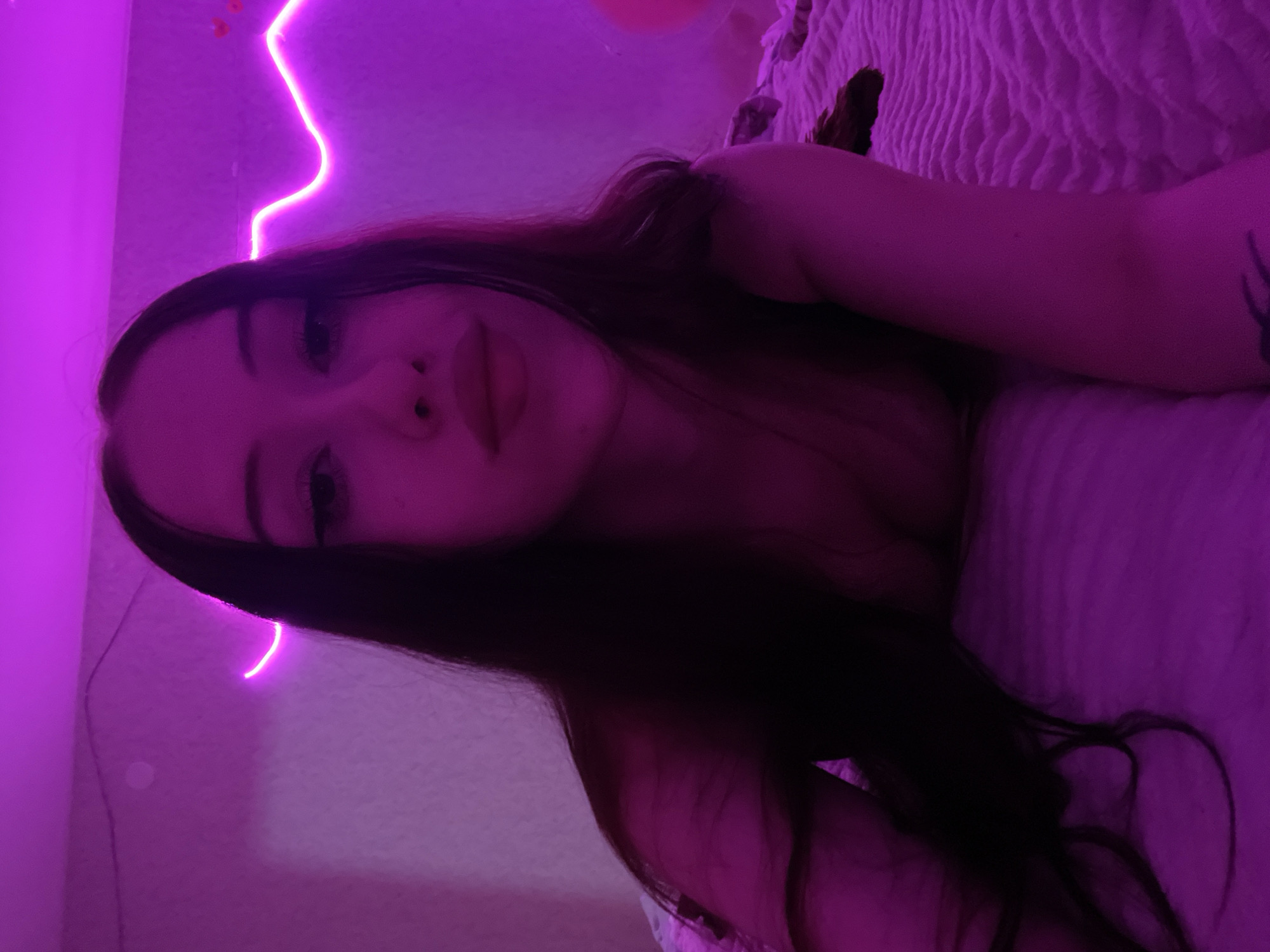 A young woman with long, dark hair is lying on a bed covered with a textured blanket. She is looking up towards the camera with her head tilted to the side. The room is illuminated by purple neon lights, which cast a strong glow on the walls and the woman's skin. The lighting creates a moody and intimate atmosphere. The woman is wearing a dark-colored top that is slightly off-shoulder, revealing a portion of her chest. A subtle tattoo is visible on her left arm. The background shows a wall with some faint decorative elements, possibly string lights. The overall scene is a selfie taken in a bedroom setting. The image does not contain any information to suggest a location in Votkinsk, Russia.