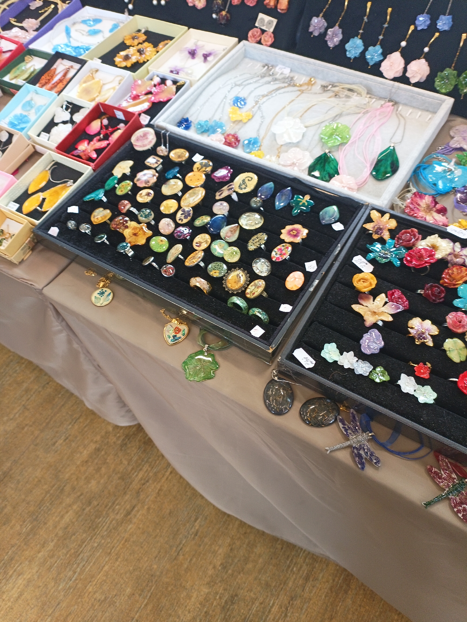 A table in a shop in Burgas, Bulgaria, displays a variety of handmade jewelry. Rings with stones, flower shapes, and dried botanical inclusions are arranged in black velvet trays. Earrings, necklaces, and pendants are also on display. The table is covered with a beige tablecloth. The floor is light brown.