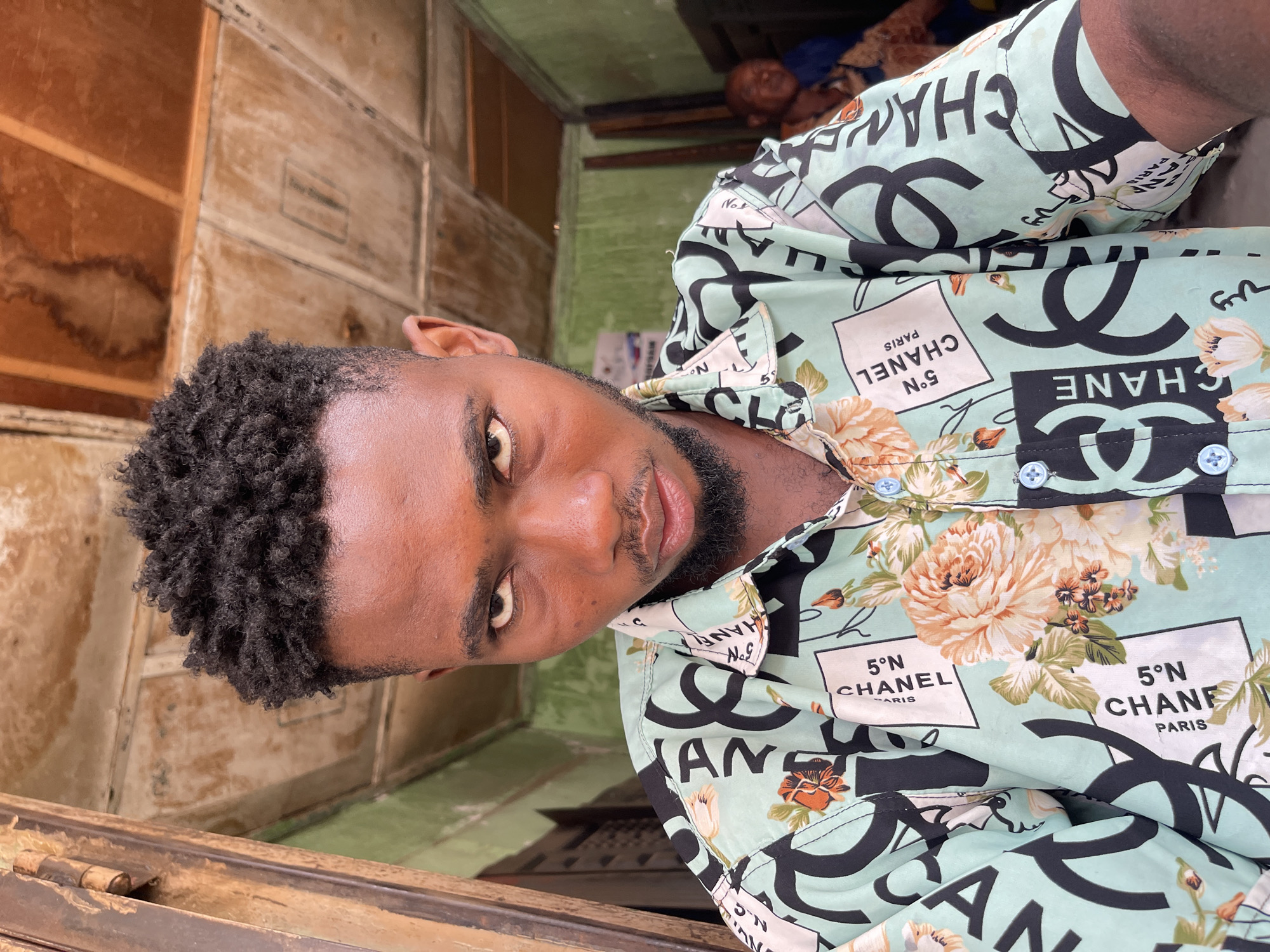 A young man with dark skin, short curly black hair, and a neatly trimmed beard looks directly at the viewer with an intense gaze. He is wearing a short-sleeved button-up shirt in a light teal or mint green color. The shirt is patterned extensively with large black interlocking 'CC' logos, reminiscent of the Chanel brand, alongside rectangular text boxes. These boxes contain phrases such as '5°N CHANEL PARIS' in black lettering, repeated throughout the fabric. Complementing the logos and text are floral designs in shades of peach, orange, and green. The shirt features light blue buttons.
The background suggests an indoor or semi-enclosed setting, likely in Zaria, Nigeria, characterized by weathered wooden panels or walls. In the upper right of the frame, another individual is partially visible, appearing to be resting or asleep, dressed in a blue garment. The man's direct gaze is a prominent feature. Visible text on the shirt includes 'PARIS,' 'CHANEL,' '5°N,' and partial words like 'CHANE' and 'CHANF' from the repeating pattern. The image is presented sideways.