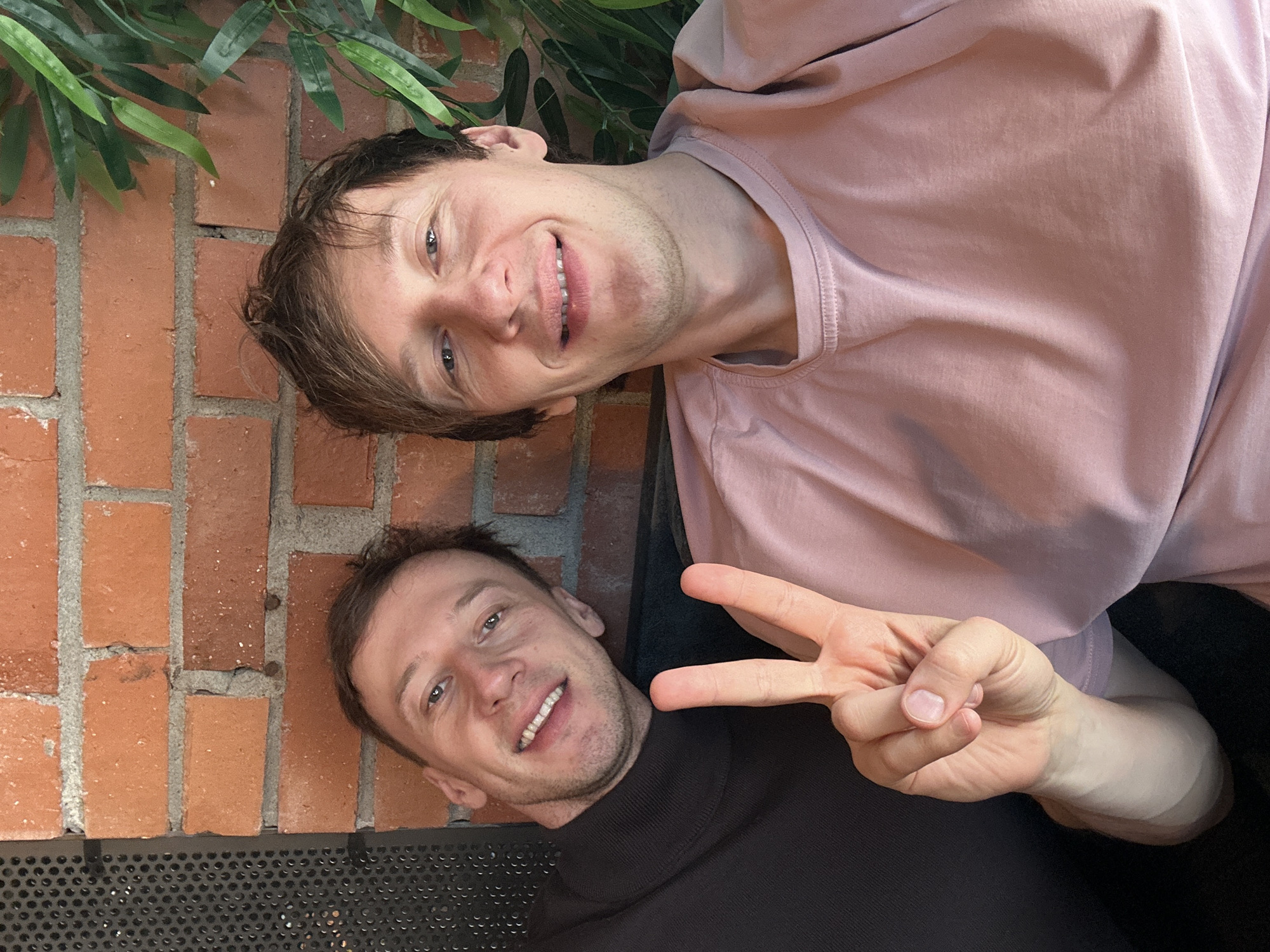 Two light-skinned men are posing for a picture outdoors. The man on the top right is wearing a light pink shirt and smiling. The man on the bottom left is wearing a black turtleneck and holding up his hand to make a peace sign. Behind them is a brick wall and some green plants. There is a grate at the bottom of the brick wall. No information indicates the location in Saint Petersburg, Russia.
