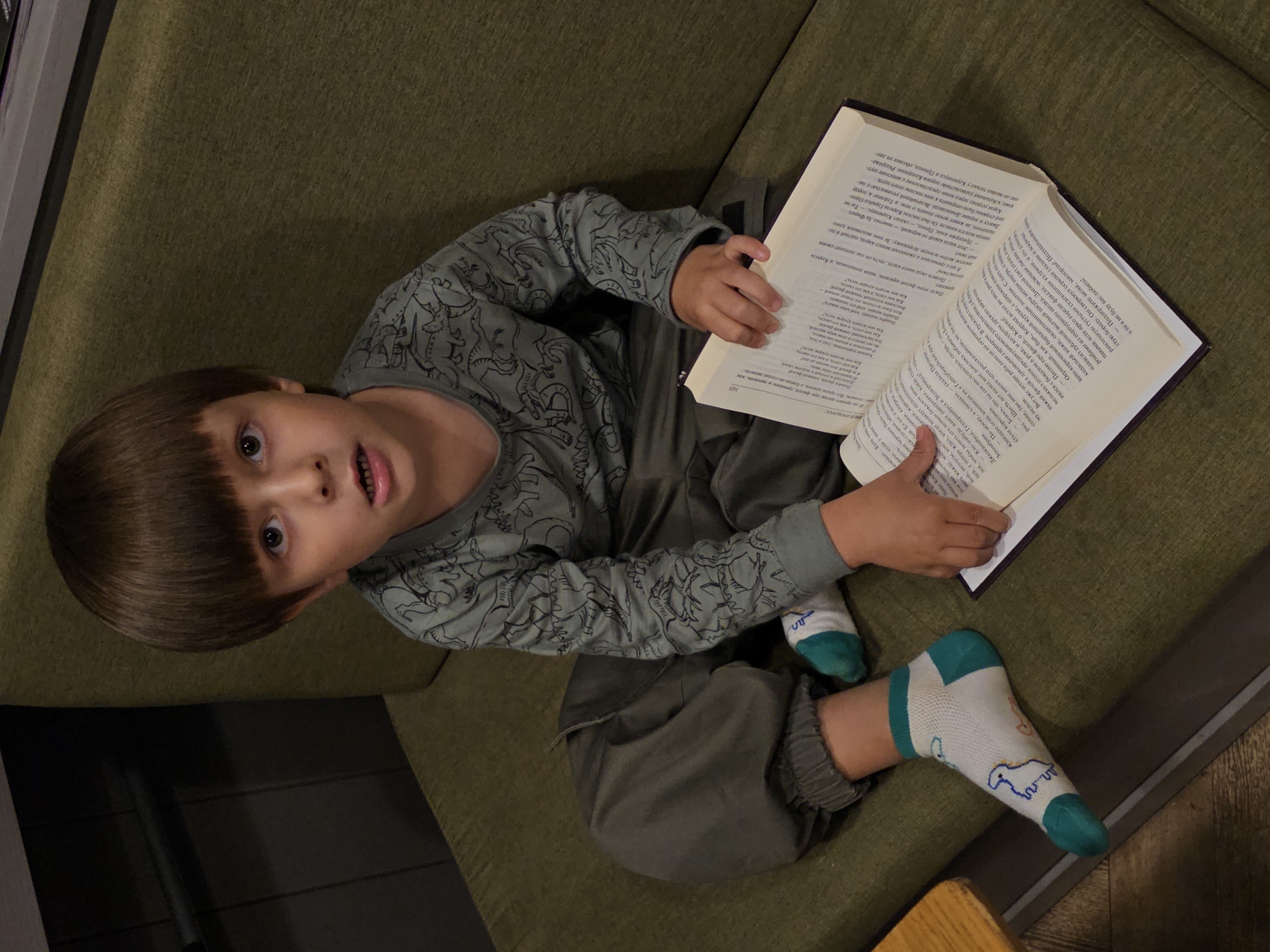 A young boy with dark, bowl-cut hair is sitting on a green upholstered bench, holding an open book. He is wearing a long-sleeved grey t-shirt with a dinosaur pattern and dark olive pants. His socks are white with green toes and heels, and feature a blue dinosaur design. The book he is holding is open to a page with text in a foreign language, and his right hand is resting on it. The boy is looking directly at the camera with a surprised expression. The environment appears to be an indoor setting, possibly a cafe or restaurant, with a dark wooden surface visible to the left of the frame. The lighting is soft and appears to be from an artificial source. The boy's location is identified as Nemchinovo, Russia.