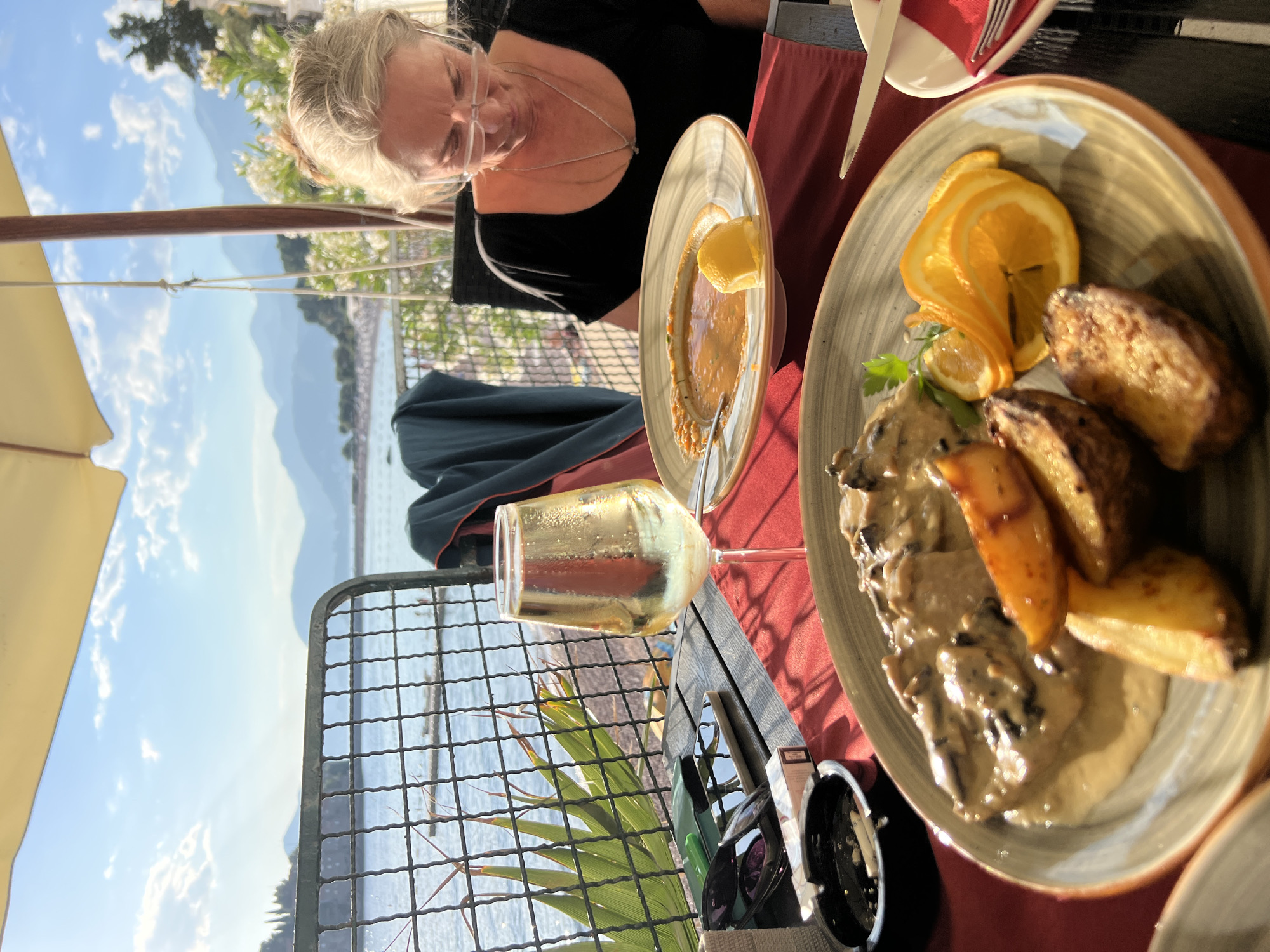 A woman with blonde hair and glasses is seated at a table outside a restaurant, with a view of the sea and mountains in the background, possibly in Sveti Stefan, Montenegro. She is looking down at a plate of food. In the foreground, there are two plates of food, one with what appears to be meat in a creamy sauce and potato wedges, and another with a soup and a lemon wedge. A glass of white wine sits on the table, along with an ashtray with cigarettes, a phone, and sunglasses. The scene is set under a large umbrella, suggesting a sunny day.