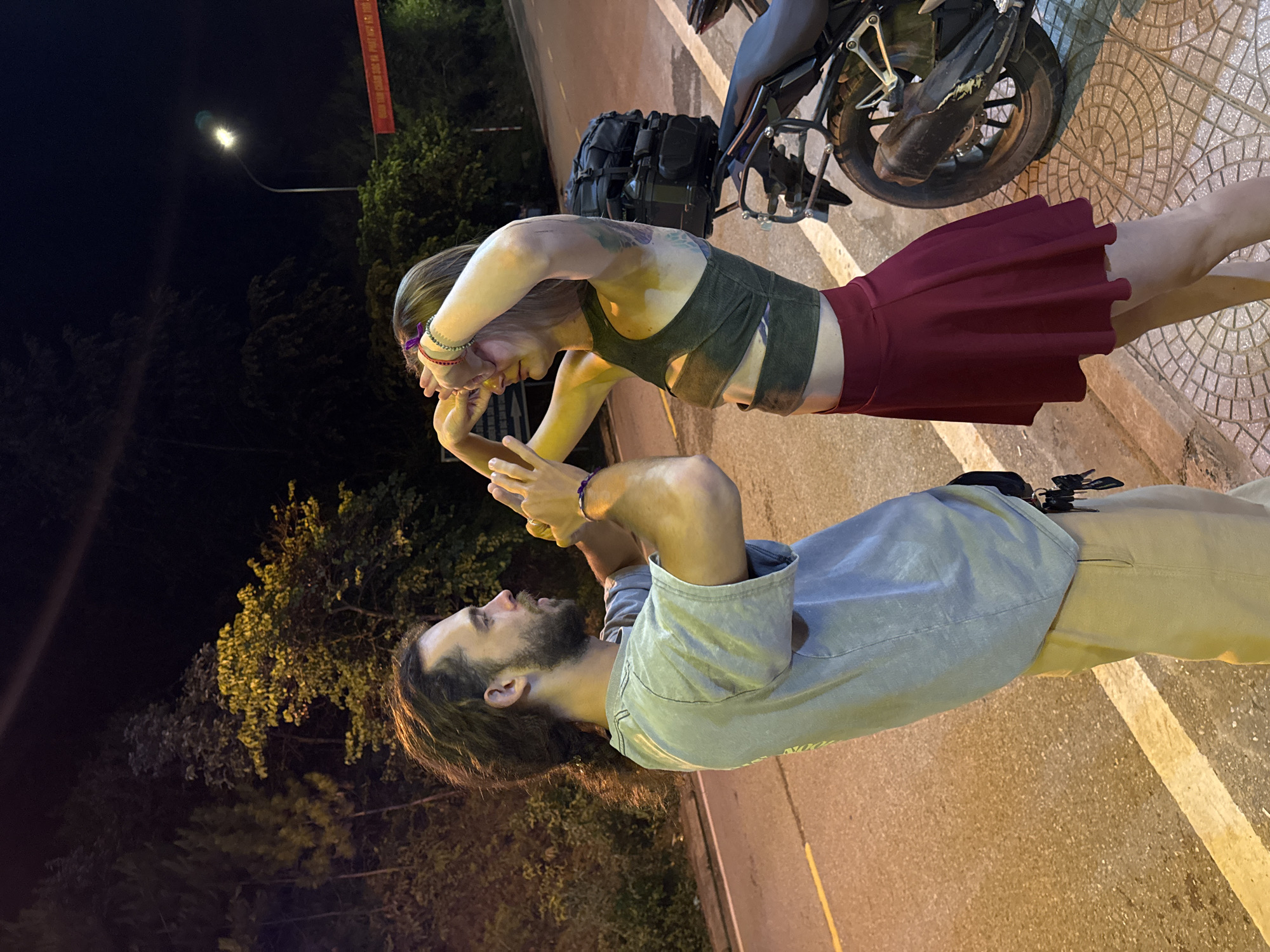 The image shows two people, a man and a woman, interacting on a roadside at night in Mù Cang Chải, Vietnam. The woman, with light-colored hair and visible arm tattoos, wears a dark green crop top and a red pleated skirt, adorned with bracelets. She leans forward, her hands near her face, appearing to look at the man or an object he holds. The man, with dark hair and a beard, is dressed in a light blue/grey long-sleeved shirt and tan pants, also wearing a bracelet. He holds a smartphone with both hands, looking towards the woman.
They are positioned beside a motorcycle, which has a dark bag or luggage strapped to its rear. The setting is illuminated by a street light directly above them, casting a bright, warm yellow light on the subjects and the immediate area. The ground consists of a patterned sidewalk next to a paved road with painted yellow lines. In the background, dark trees are silhouetted against the night sky, with another street light visible in the distance. A red banner with indecipherable Vietnamese text hangs from a pole further back. The scene suggests a casual, intimate moment during a stop on a journey at night.