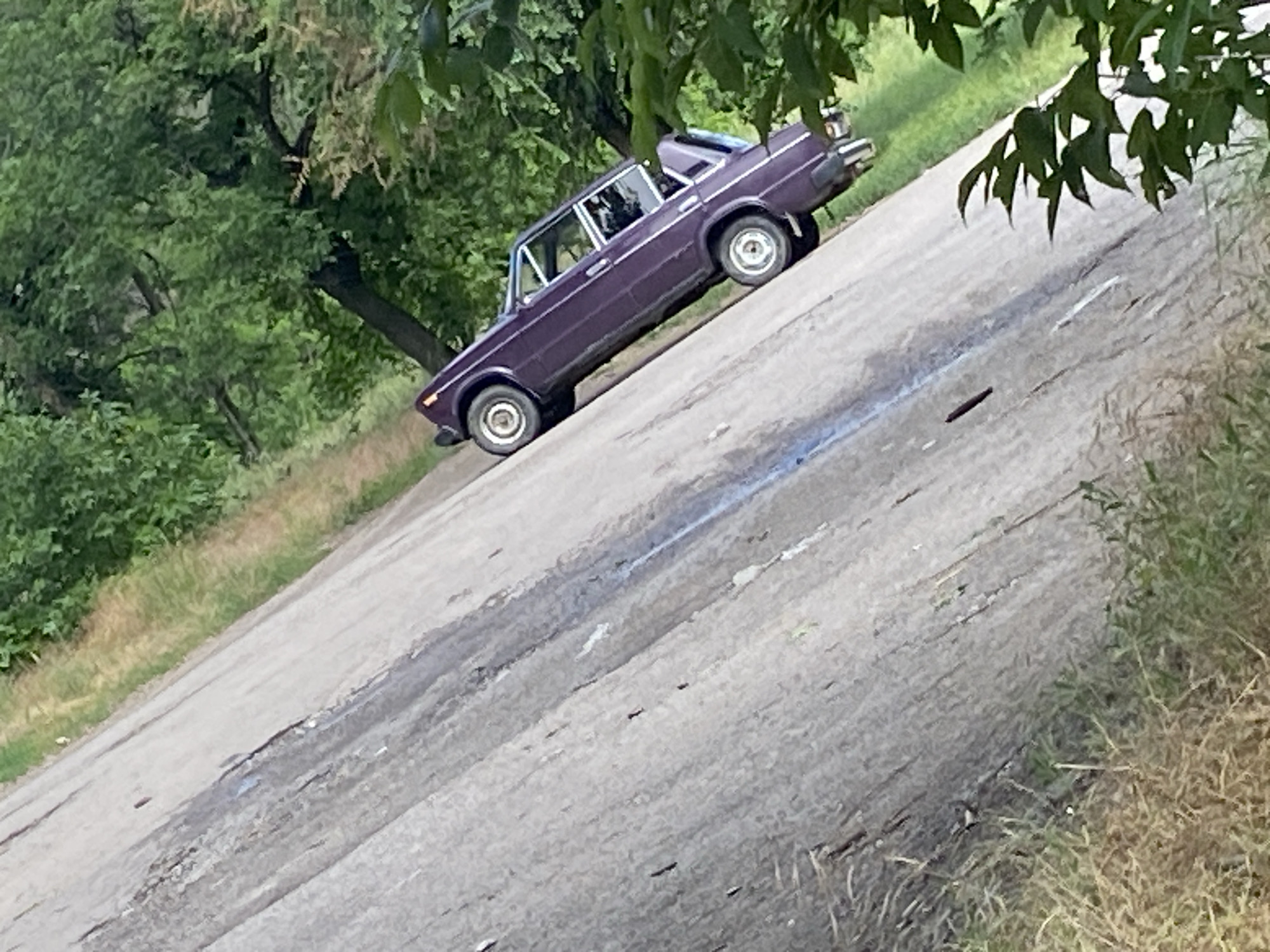 An old, dark purple sedan is parked on the side of a cracked asphalt road, angled upwards to the right. The car has visible damage to its bumper. The road is bordered by dry grass and green foliage on the left and lush green trees on the right. The scene appears to be in a rural or undeveloped area.