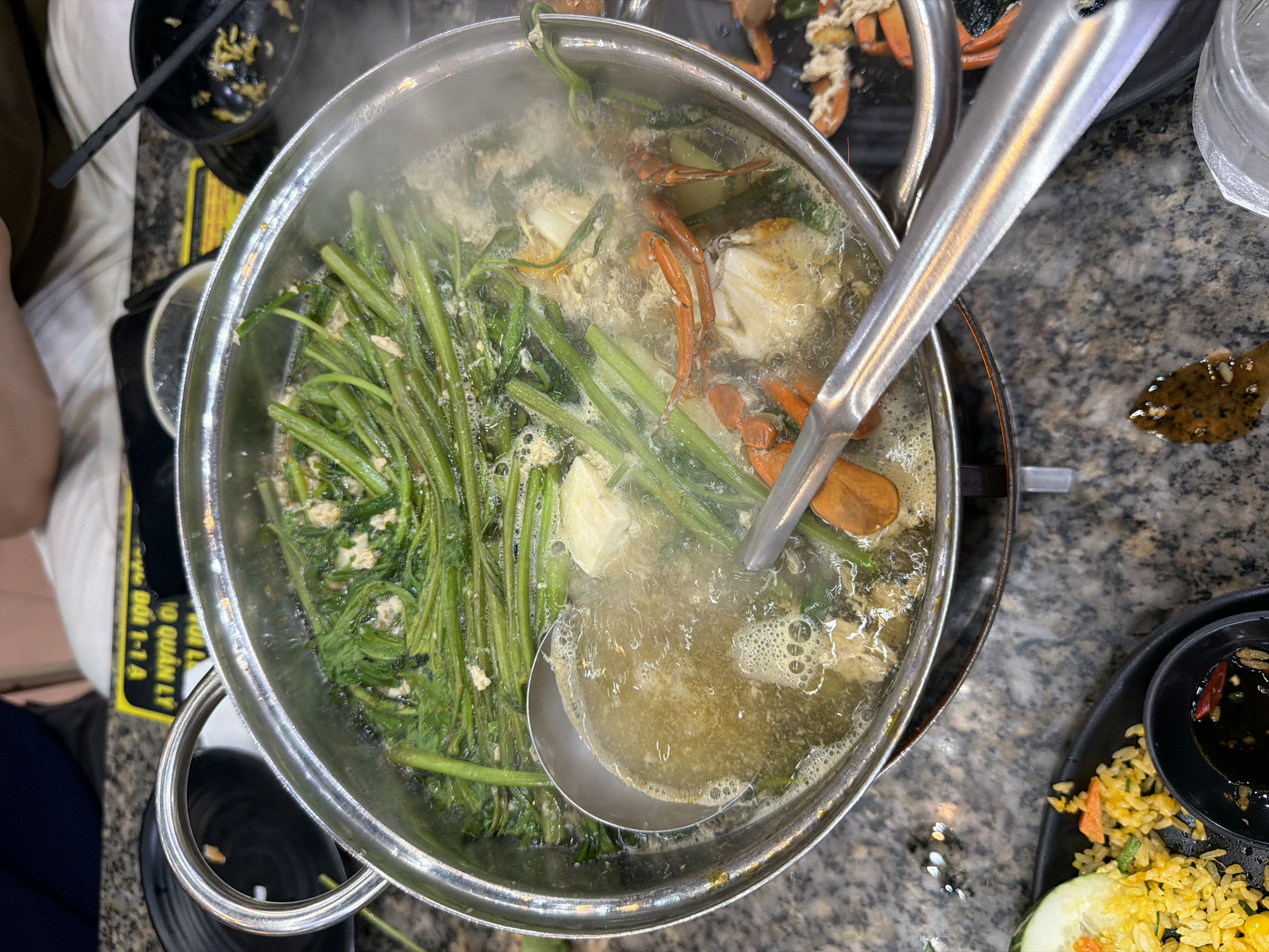 An overhead shot captures a vibrant hot pot meal in a dining setting in Quy Nhon Ward, Vietnam. The central focus is a large, circular metal hot pot filled with bubbling soup, emitting steam. Inside the pot, numerous long, green leafy vegetables (resembling water spinach or morning glory) are submerged, alongside several pieces of cooked crab, identifiable by their orange shells and white meat. Some lighter-colored, possibly tofu or egg, pieces are also visible in the broth. A large metal ladle rests inside the pot, ready for serving.
To the left, a black bowl containing chopsticks and some food remnants, possibly noodles, is visible on a black and yellow electric hot plate beneath the pot. Partially visible on the far left is the arm or leg of a person seated at the table. On the right side of the image, a black plate holds a serving of yellow fried rice, a slice of cucumber, and a small black bowl filled with a dark dipping sauce, garnished with red chilies and spices. A small spill of dark liquid is present on the speckled gray granite table surface near this plate. In the upper right corner, clear plastic cups are stacked.
The scene depicts a communal dining experience, with the hot pot as the main event, suggesting a freshly cooked and shared meal. The bubbling soup and steam emphasize the heat of the dish. A yellow sign beneath the hot pot is partially visible, showing Vietnamese text including "...O QUẢN LÝ..." (management) and "...ĐỐI 1-1-A".