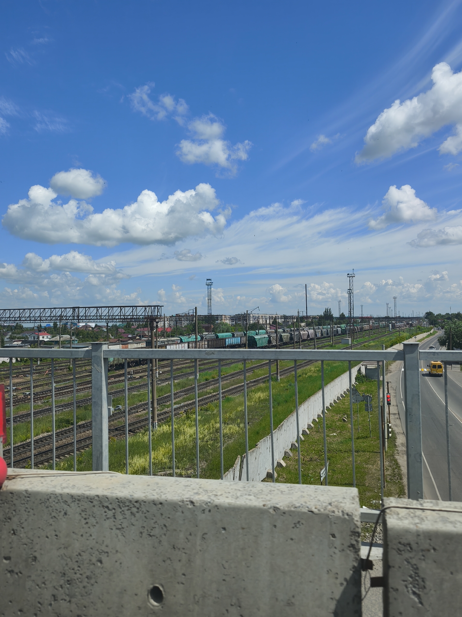 The image depicts a view from an elevated position, likely a bridge or overpass, overlooking a railway yard and a road in the city of Kanash, Russia. The foreground is dominated by a concrete barrier, suggesting a pedestrian walkway or elevated thoroughfare. Beyond the barrier, a metal railing separates the viewer from the scene below, which consists of numerous parallel train tracks crisscrossing a grassy area. Several freight trains, loaded with what appear to be cylindrical containers, are visible on these tracks, stretching into the distance.
To the right of the railway yard, a paved road runs parallel, with a yellow vehicle, possibly a bus or van, driving on it. A traffic light with a red signal is also visible on the side of the road. In the background, a cityscape with houses and industrial buildings can be seen, punctuated by tall utility poles. The sky above is a clear blue, dotted with fluffy white clouds, indicating fair weather. The overall impression is of an active industrial and transportation hub on a bright, sunny day.