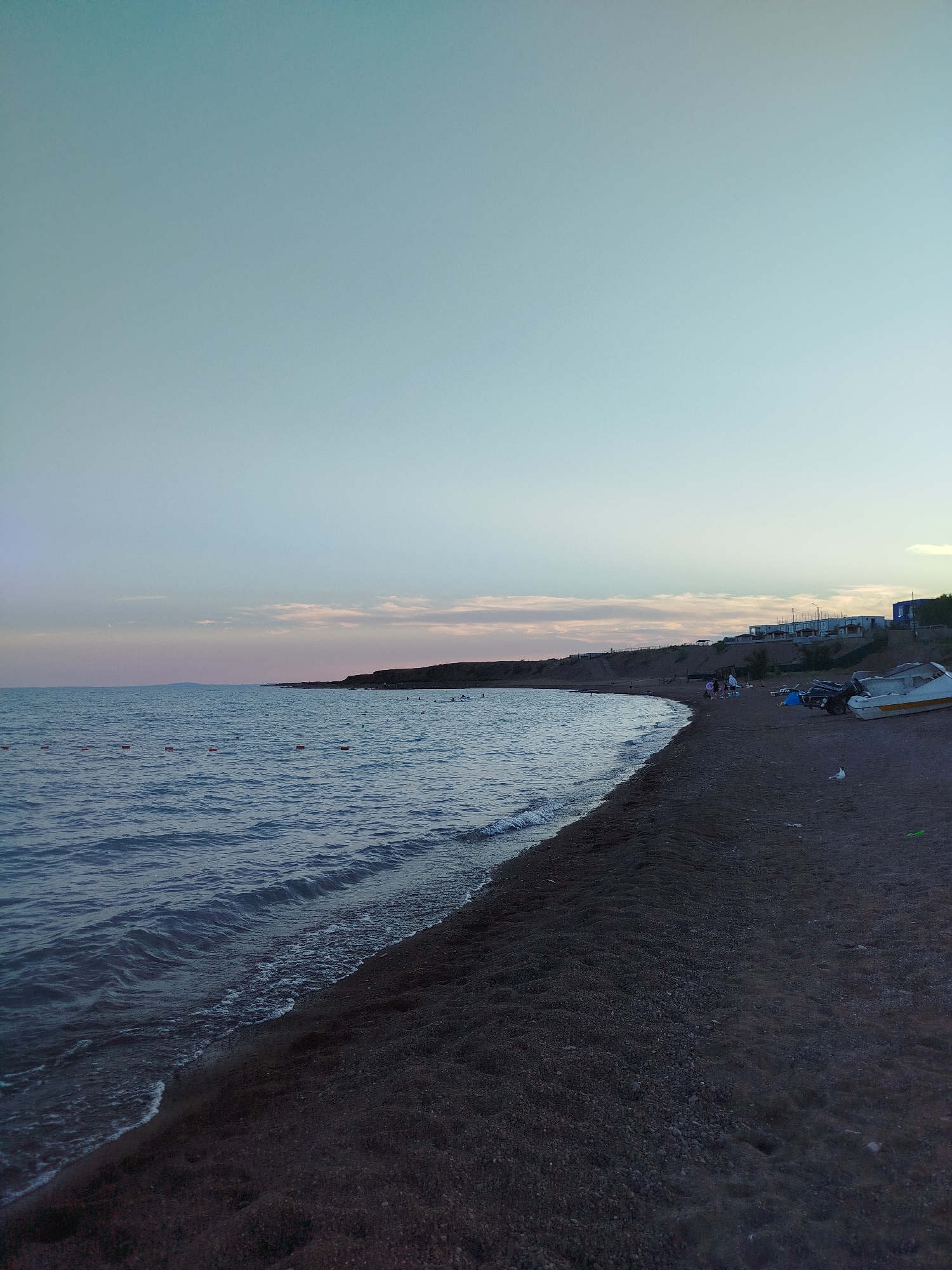The image shows a dark, pebbly beach beside a calm, dark blue sea. The sky is clear and a pale gradient of blue and pink, suggesting either dawn or dusk. Along the shoreline, small orange buoys are visible in the water, marking a designated area. Further back, several people are scattered on the beach, some sitting, and a few are in the water. A small cluster of buildings, likely residences or resort accommodations, are situated on the elevated land behind the beach. Two small boats are parked on the sand near the buildings. A lone white seagull is perched on the beach.
