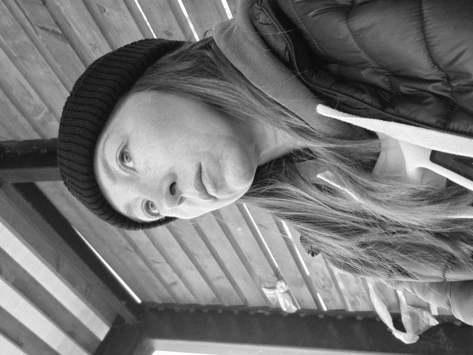 A grayscale, angled close-up captures a woman with light hair wearing a dark, possibly knitted beanie and a puffy jacket with a hoodie visible underneath. The background is a wooden structure, possibly a pergola or a similar open-air shelter, with sunlight creating alternating stripes of light and shadow. The woman's face is the main focus, with her eyes looking towards the upper left of the frame. The location is potentially in the city of Chaikovsky, Russia, inferred from the request. There is a hint of something white hanging from the jacket. The shot appears to be taken from a slightly low angle.