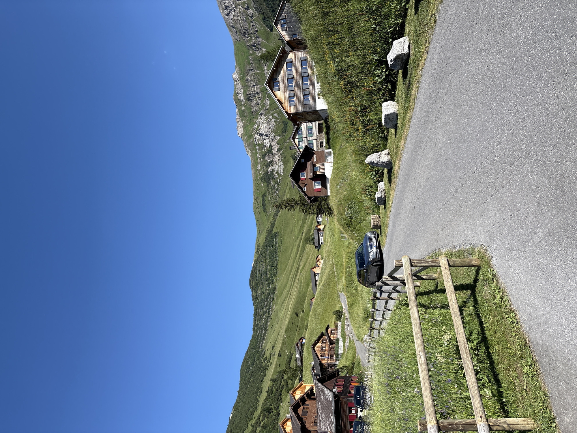 The image depicts a scenic view of Malbun, Liechtenstein, characterized by traditional wooden chalets nestled on lush green hillsides under a clear blue sky. A paved road winds through the village, with a wooden fence bordering one side, separating it from a field of wildflowers and tall grass. A silver station wagon is parked on the right side of the road, and other vehicles are visible parked near the chalets on the left. The chalets themselves are multi-story structures with wooden facades and dark-shuttered windows, typical of alpine architecture. The background features gently sloping, verdant mountains, with some rocky outcrops visible higher up. The overall impression is one of tranquility and natural beauty, typical of a mountain resort town in the summer.