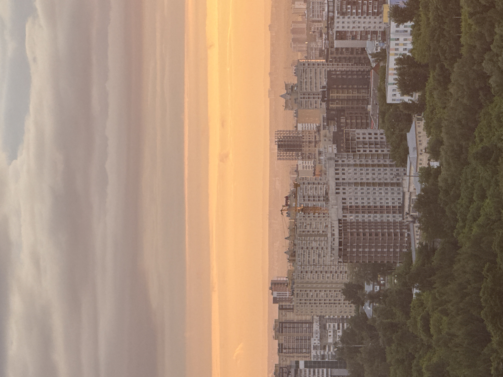 This image presents an expansive, elevated view of the city of Гамово, Russia, captured during either sunrise or sunset. The upper sky displays layered, pale greyish-pink clouds. Below this, a distinct horizontal band of intense warm orange-yellow light dominates the mid-sky. A hazy, distant landmass or urban outline is faintly visible beneath this bright horizon.
The mid-ground is densely occupied by numerous multi-story buildings, appearing largely residential or mixed-use. Multiple structures are visibly under construction, characterized by exposed concrete frameworks, unfinished facades, and the presence of at least one yellow-orange construction crane on the right side of the frame. The buildings exhibit varying architectural styles, predominantly functional and modern.
The foreground is dominated by a substantial, dense expanse of dark green trees, forming a forest or extensive parkland. Several smaller, lower-rise buildings are situated within or at the immediate edge of this forested area. The overall scene is characterized by soft, diffuse lighting and a pervasive atmospheric haze, with no strong, defined shadows cast by individual structures. No people or specific activities are discernible.