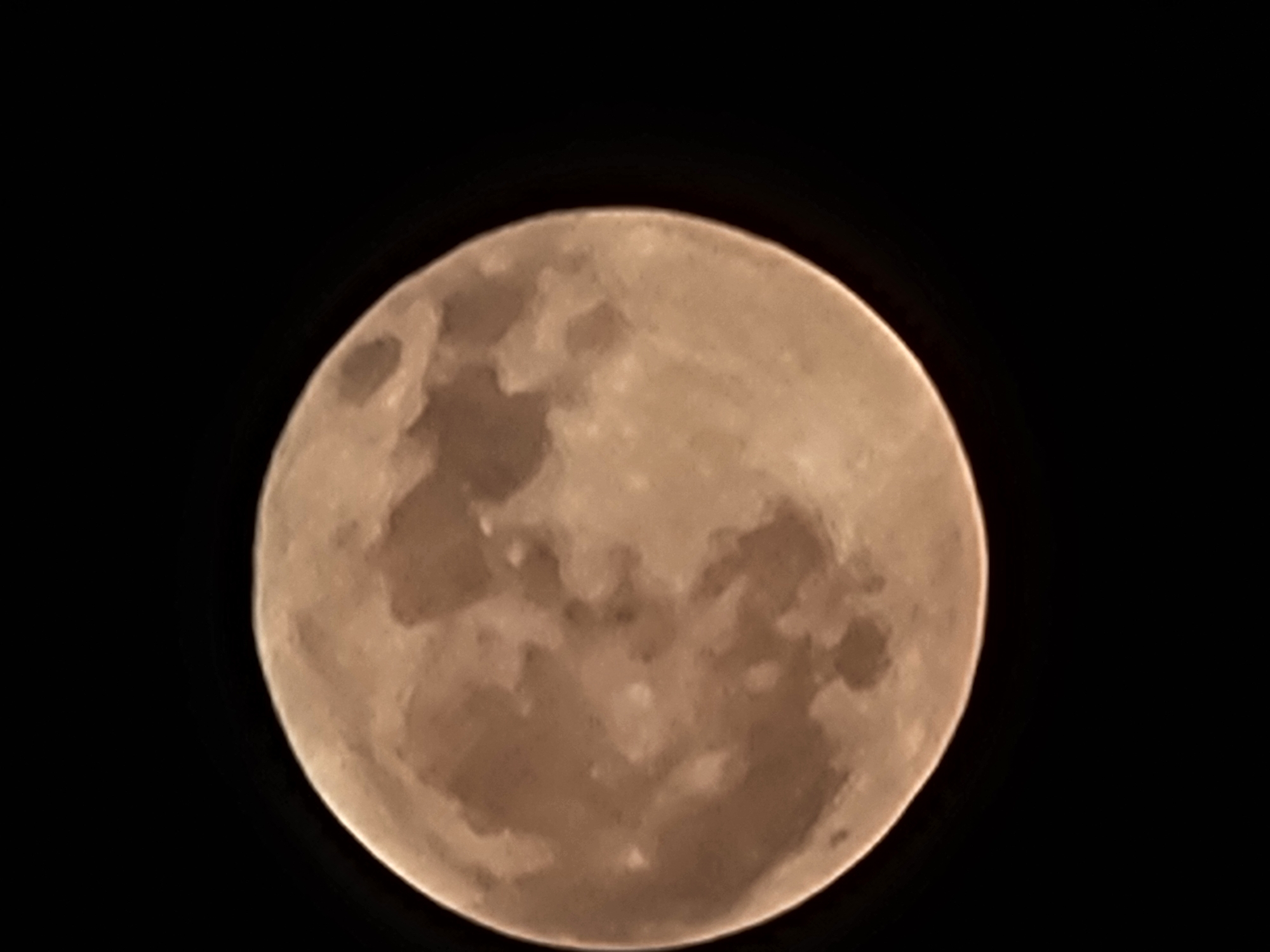 A close-up, low-angle, full-body shot of a person's face, framed by the dark sky. The person's face is illuminated by the soft, warm glow of the full moon, which occupies the majority of the frame. The moon's surface is visible, with craters and maria creating a textured, detailed appearance. The person's features are indistinct due to the low light conditions and focus on the moon. The background is entirely black, emphasizing the celestial subject. There are no discernible elements that place this image in Quarteira, Portugal.
