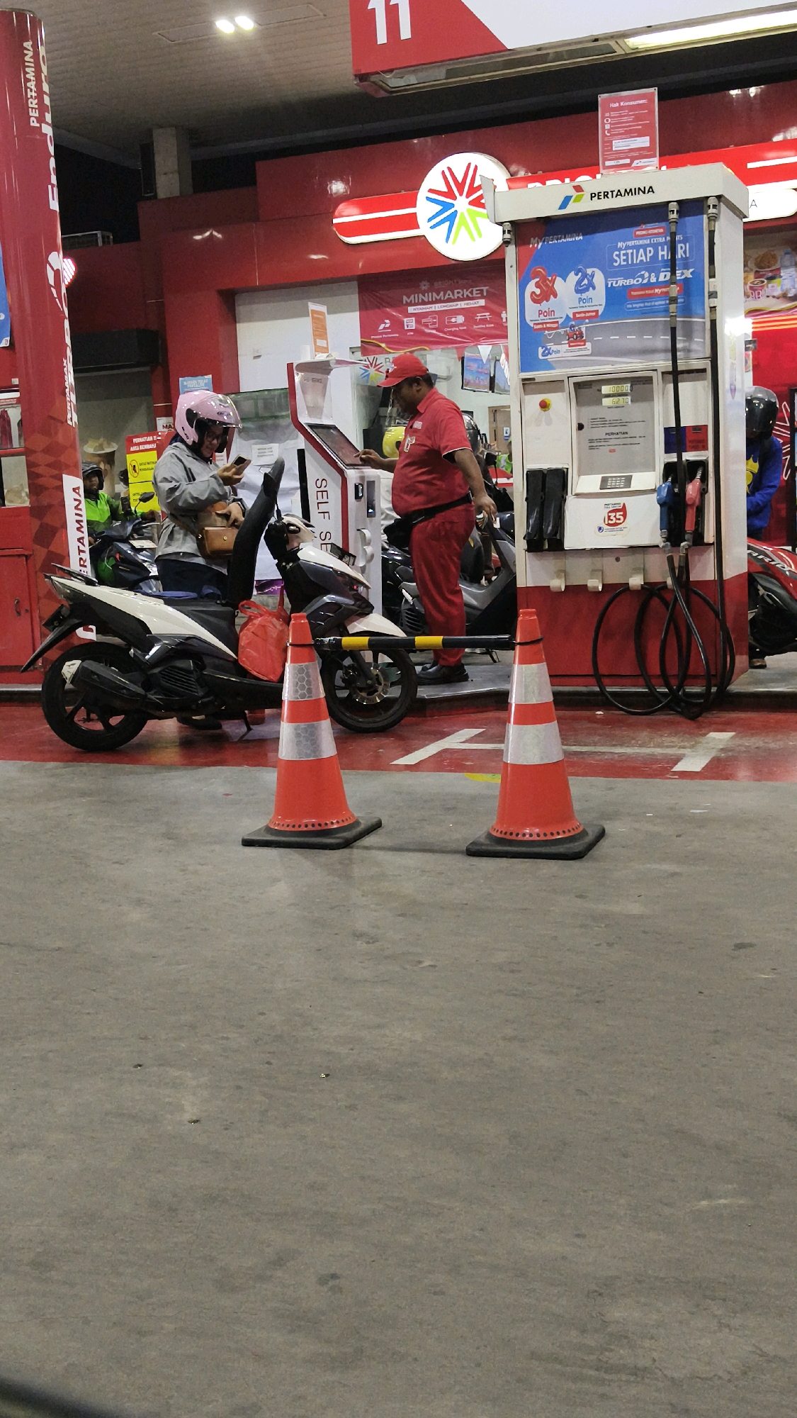 At a Pertamina gas station in Central Jakarta, Indonesia, a person wearing a pink helmet and a gray jacket sits on a white scooter, looking at their phone. To their right, a gas station attendant in a red uniform stands at a self-service payment kiosk, facing away from the viewer and interacting with the screen. The kiosk is positioned next to a gas pump. Two orange traffic cones with reflective white bands are placed on the red-and-white marked concrete floor in the foreground. In the background, a minimart sign is visible, along with other gas pumps and vehicles. The lighting appears to be artificial, suggesting it is nighttime or an indoor covered area.