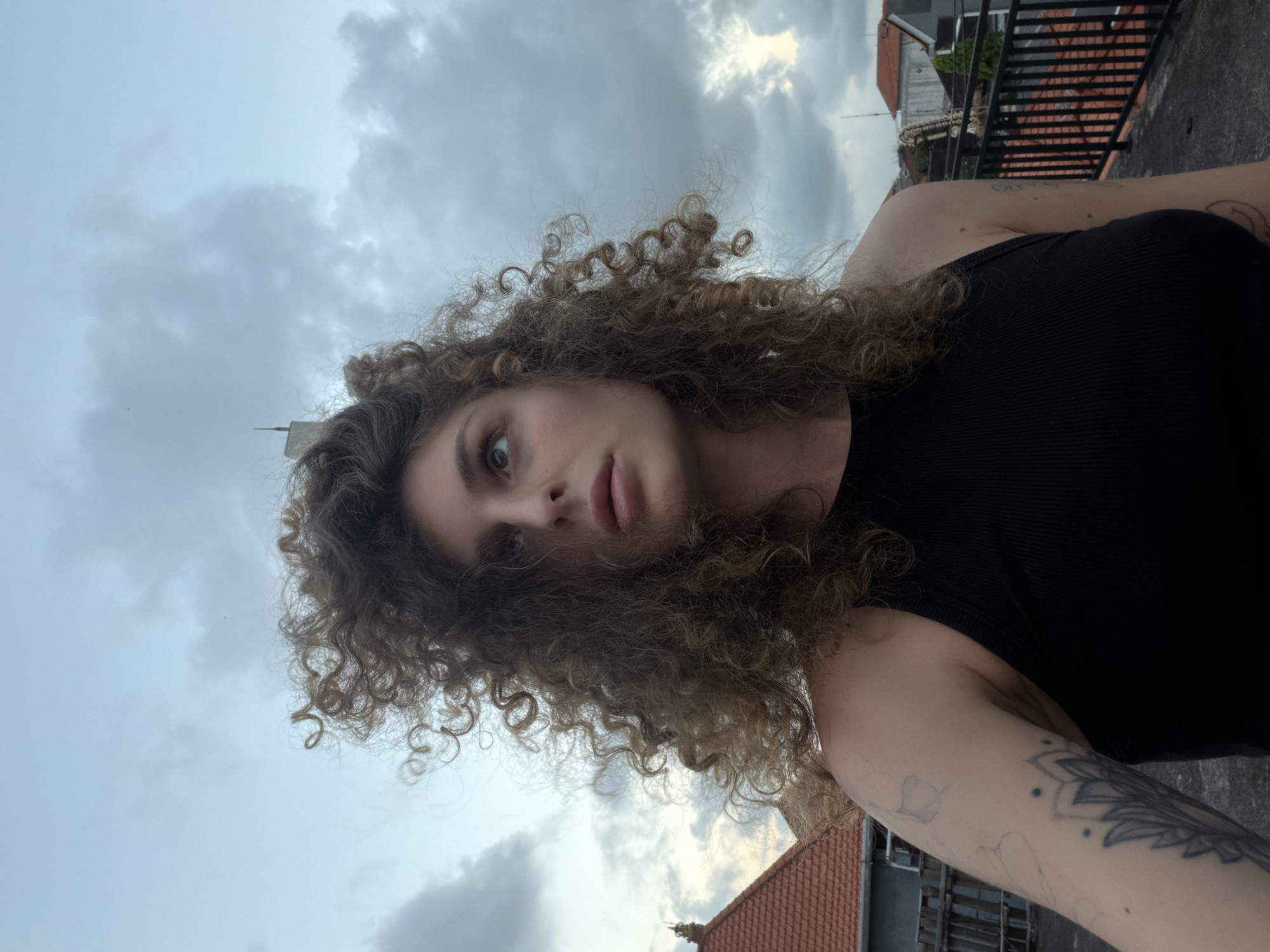 A woman with curly brown hair stands on a rooftop in North Kuta, Indonesia, looking up and to the right. She is wearing a black tank top. Her left arm has tattoos. The sky is cloudy, and red-tiled roofs of buildings are visible in the background, along with a metal railing.