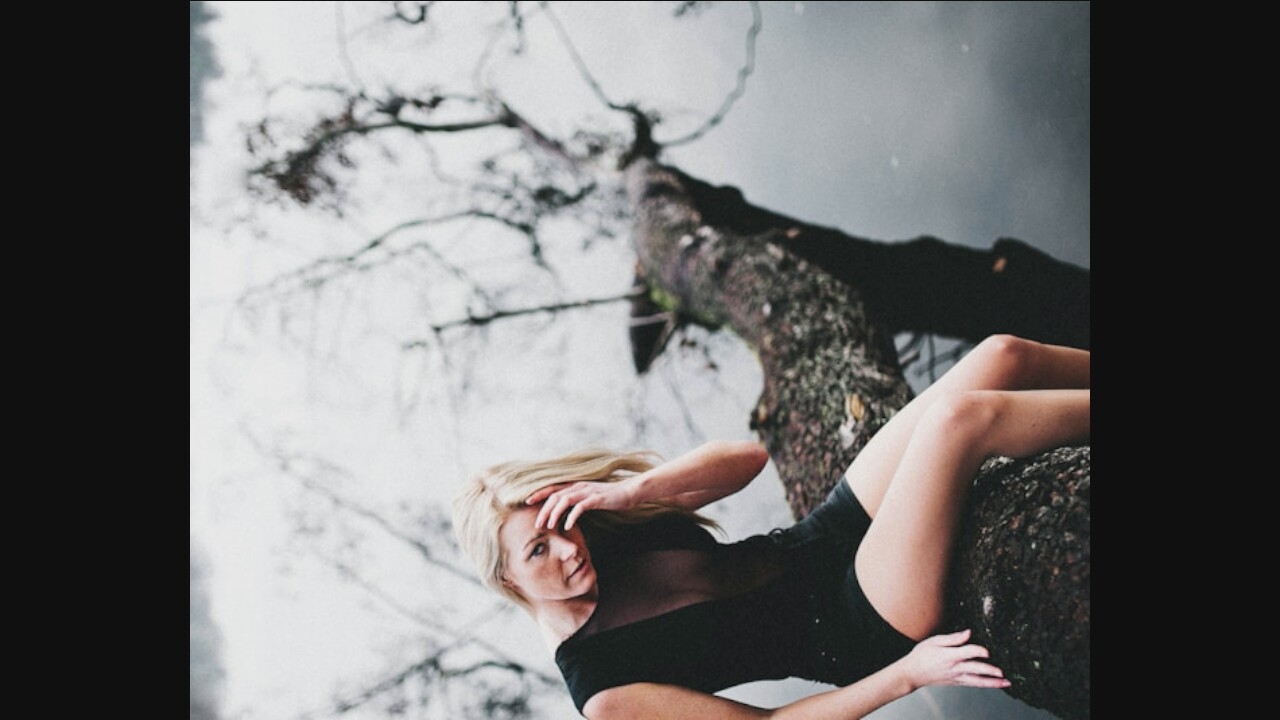 A blonde woman is sitting on a fallen tree that is partially submerged in water. She is wearing a black, sleeveless dress with a sheer neckline. Her hand is resting on her forehead, and she is looking down. The background is blurry, showing leafless trees and a body of water. The overall scene is somber and moody.